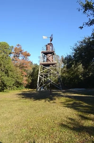 A wooden windmill with a metal rotor on a grassy field surrounded by trees under a clear blue sky.