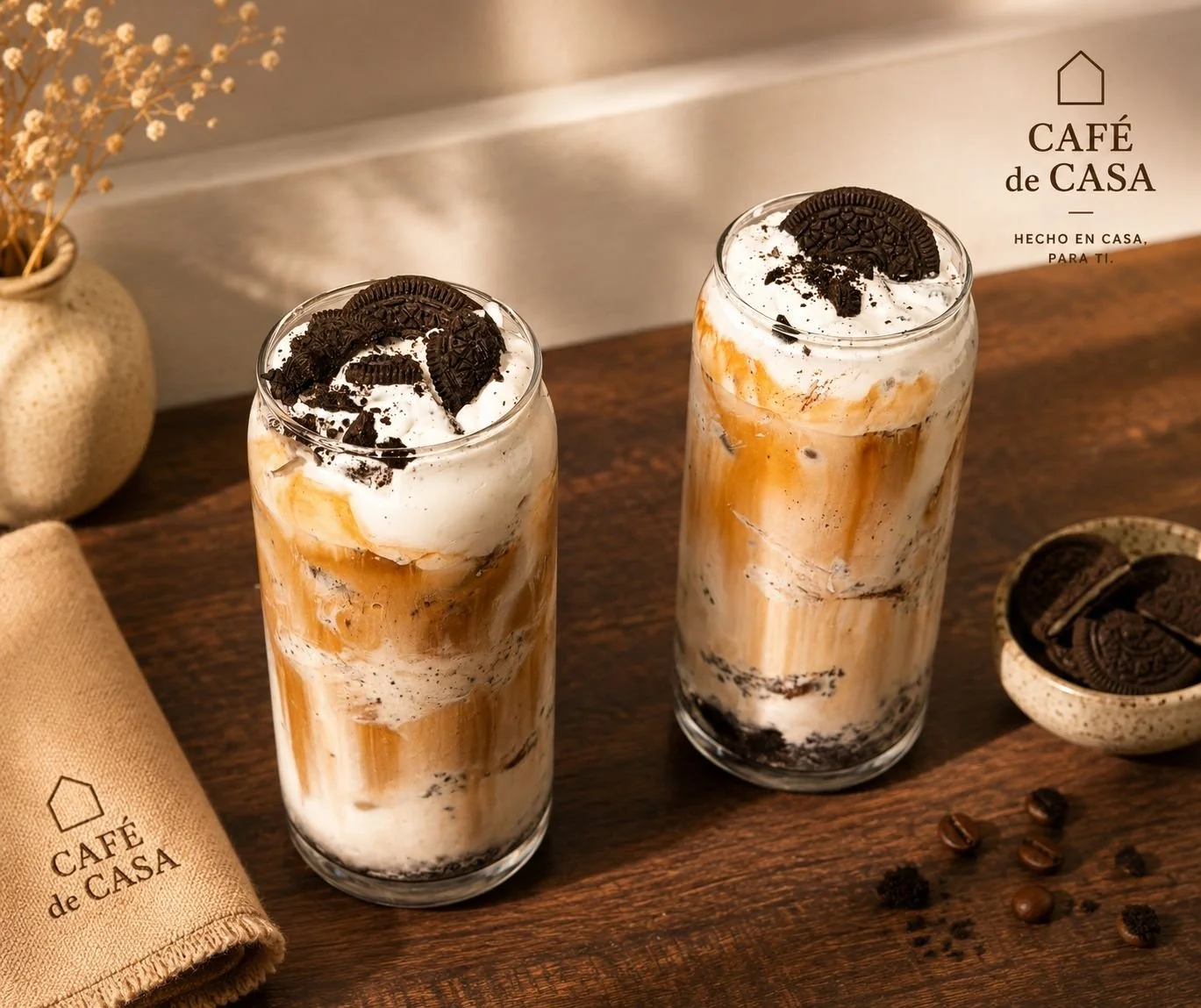 Two tall glasses of Oreo caramel frappuccino on a wooden surface, topped with whipped cream and Oreo cookies, with a small bowl of Oreo cookies and scattered coffee beans nearby.