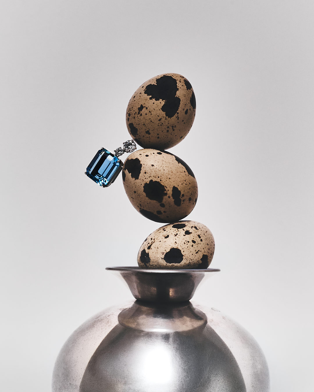 Tres huevos de codorniz apilados uno sobre otro, con un anillo de compromiso con un gran diamante y una gema azul encima del huevo superior, sobre una superficie metálica.