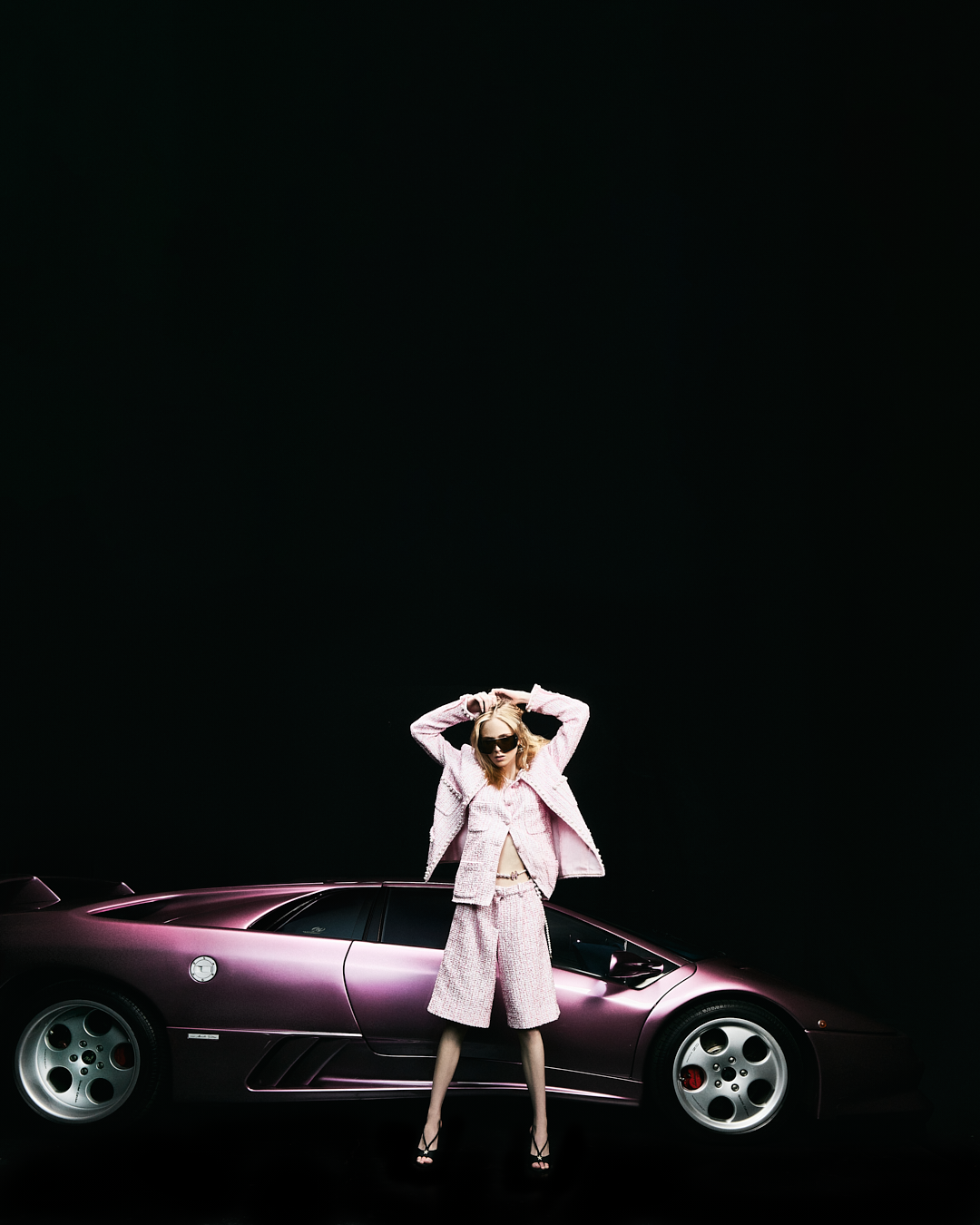 Mujer con gafas de sol, ropa rosa, posando frente a un coche deportivo de color rosa metálico, en un fondo negro.