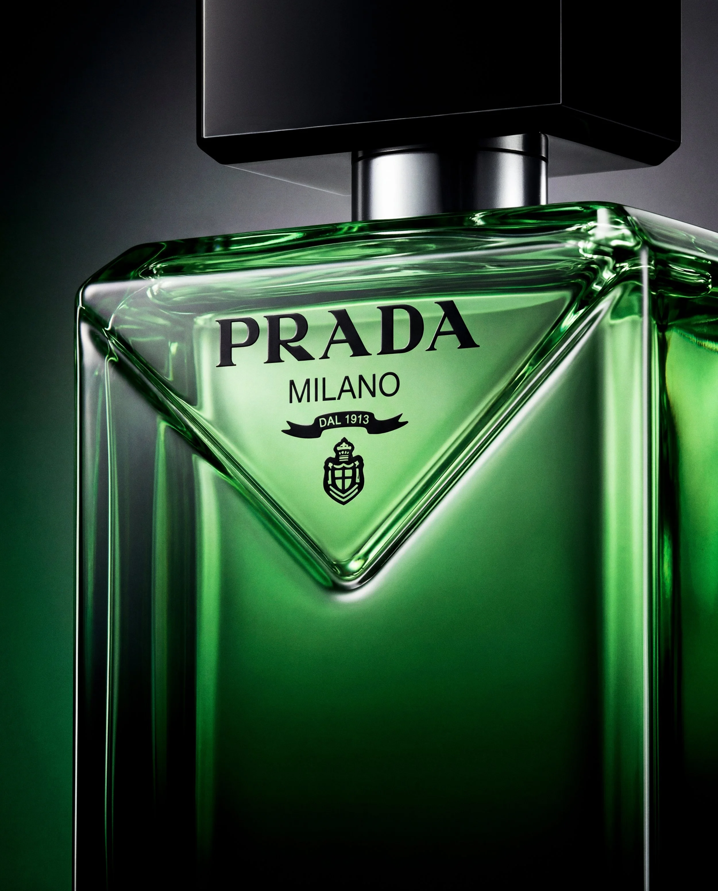 Frasco de perfume verde de Prada con etiqueta que dice 'Prada Milano' y un escudo, en un fondo gris oscuro.