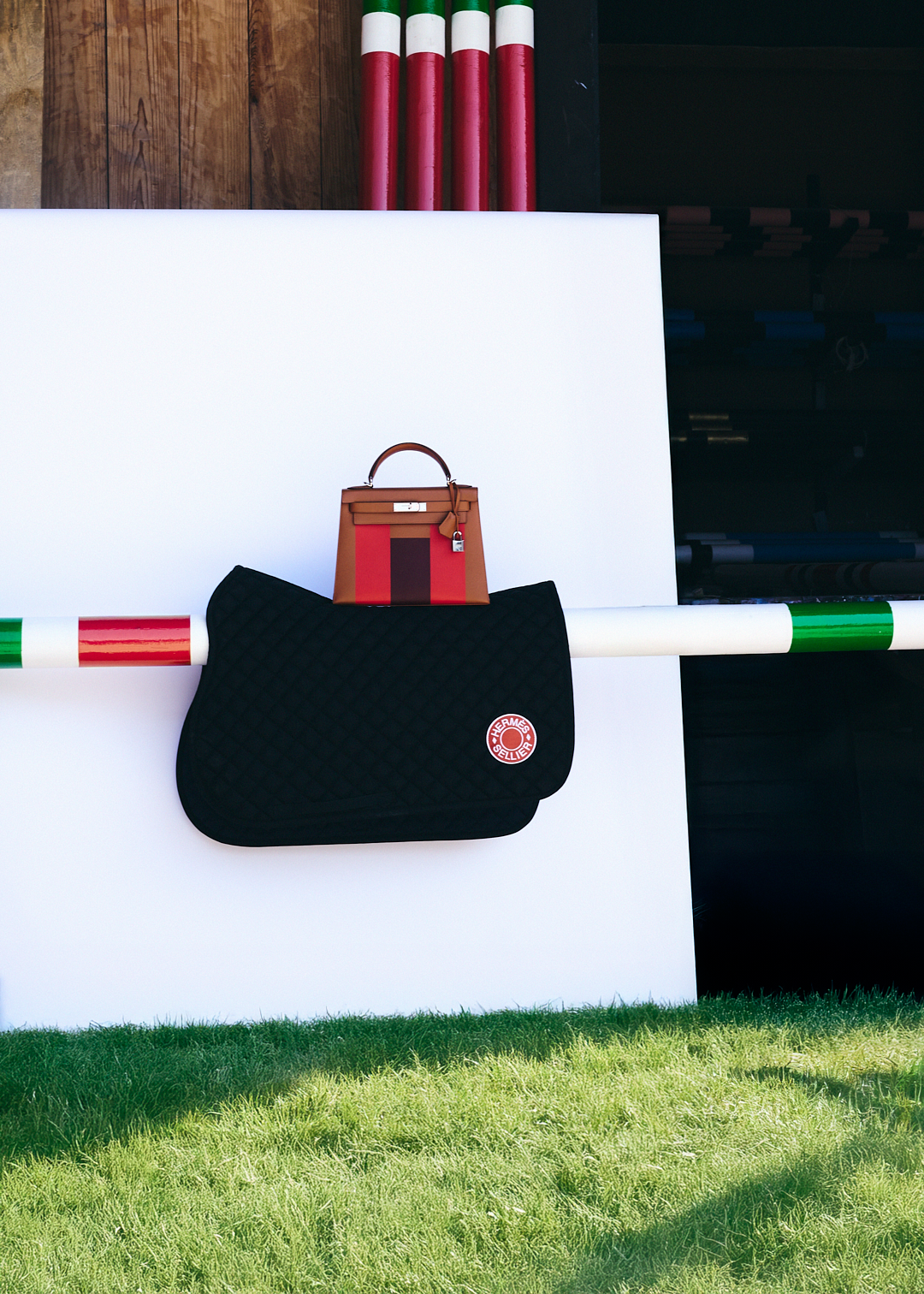 Un bolso color madera y rojo y una bolsa negra con el logotipo de Hermes en una estructura blanca con pasto verde en el suelo.