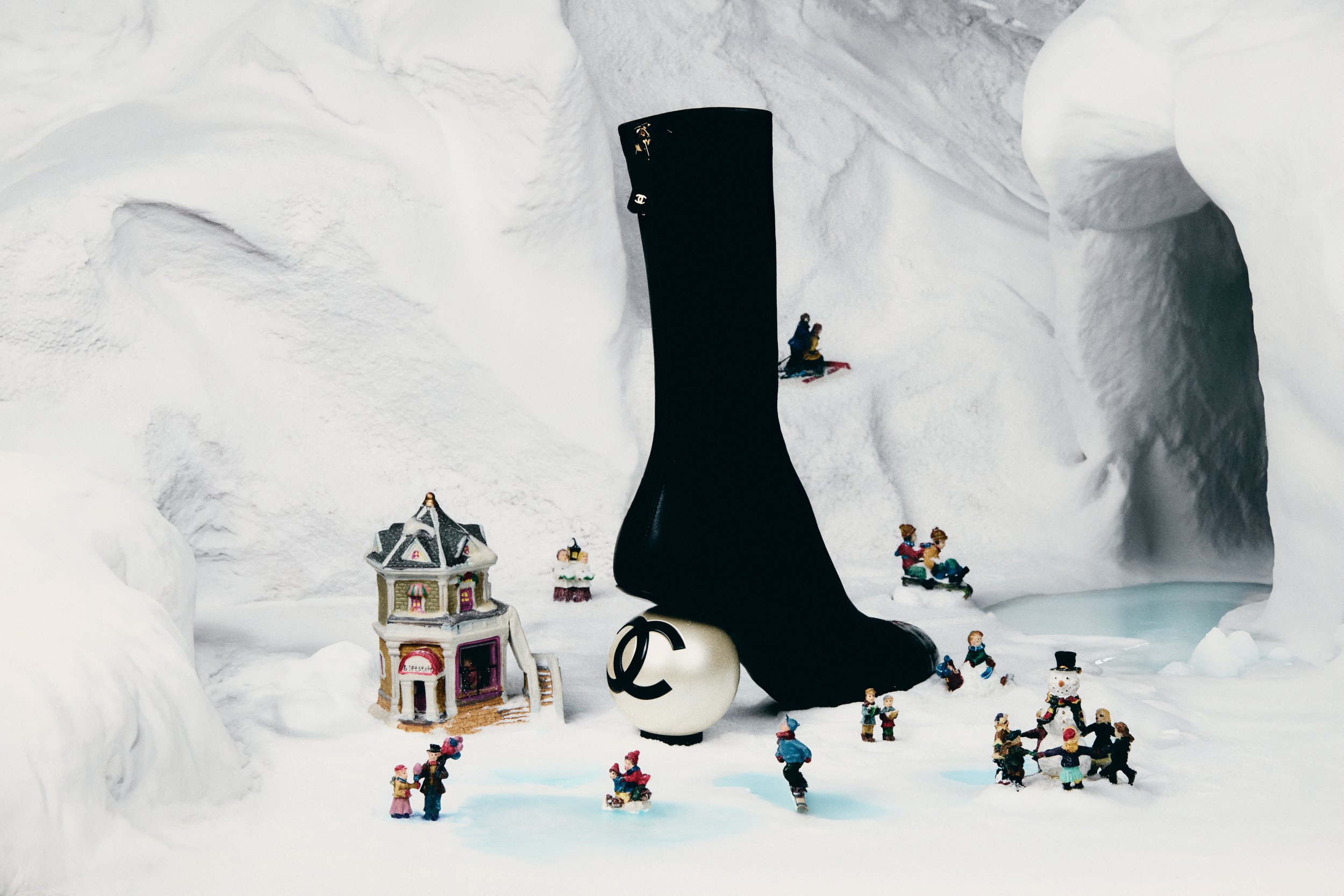 Escena invernal con figuras de Navidad y muñecos, incluyendo un calcetín negro con logotipo de Chanel y una figura del símbolo del dólar, rodeados de nieve y mini casas decoradas.