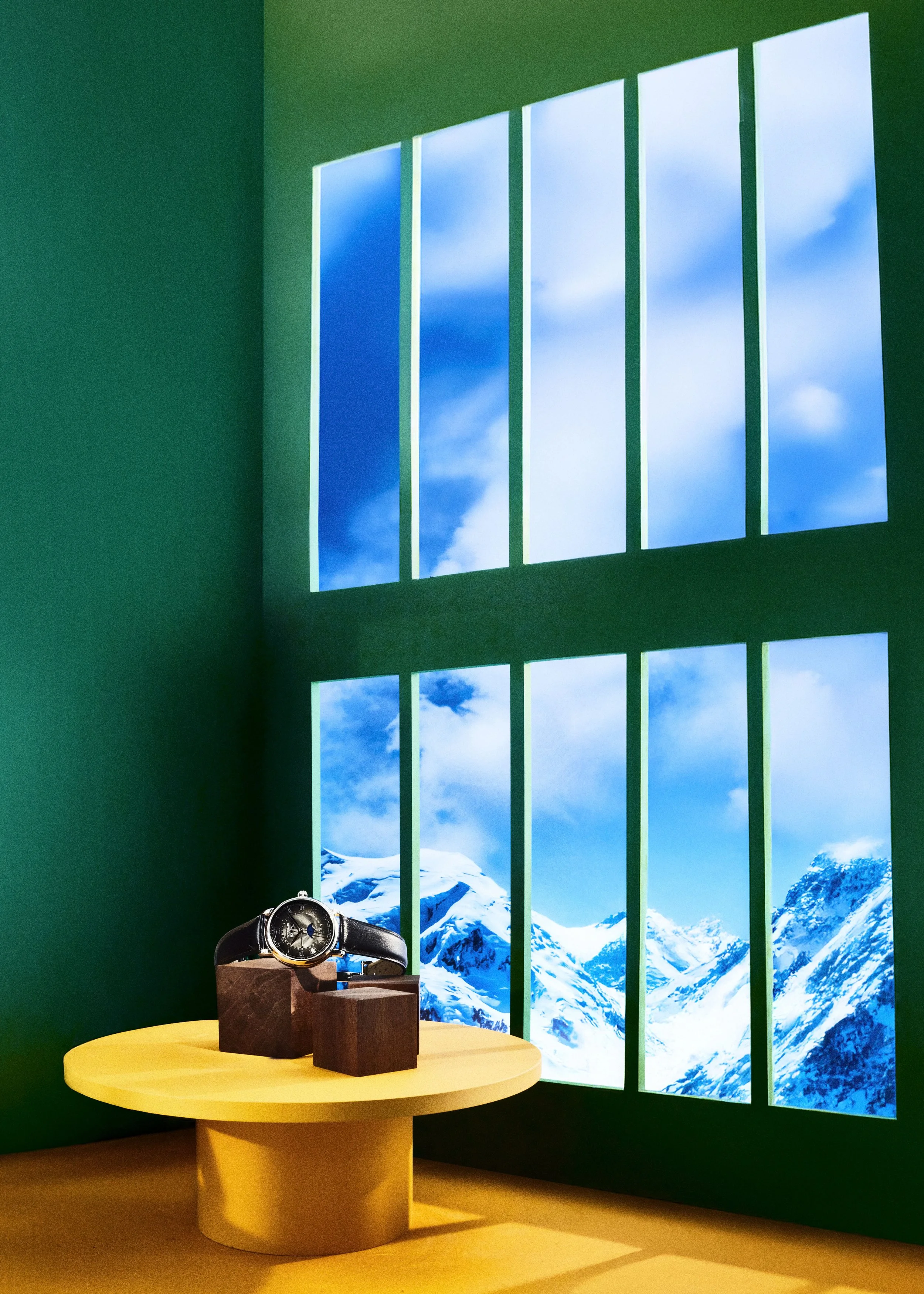 Reloj de pulsera negro en exhibición sobre una mesa amarilla, con un paisaje montañoso nevado y un cielo azul visible a través de ventanas grandes.