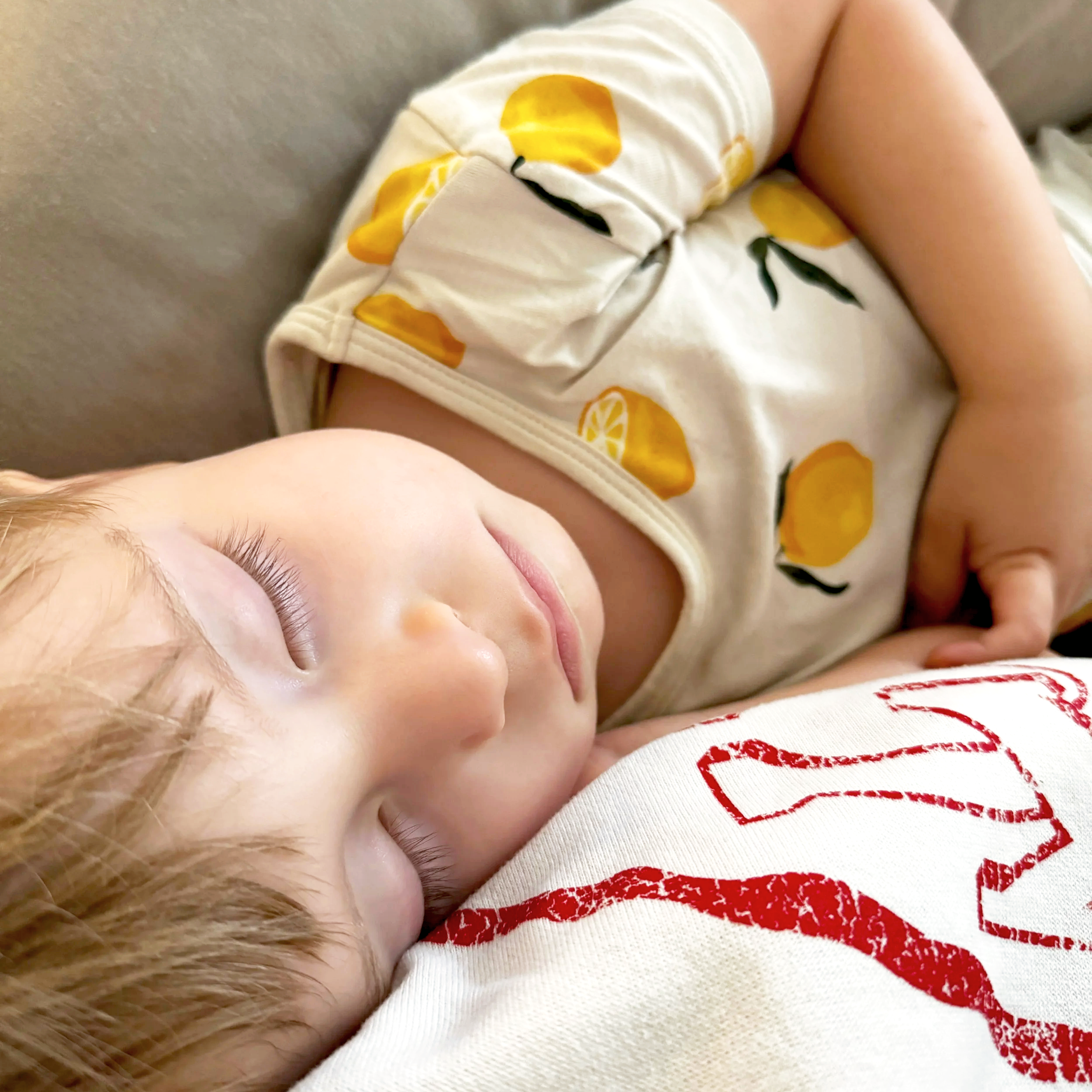 Close-up of a sleeping baby wearing a white onesie with a lemon print, resting on a soft surface.