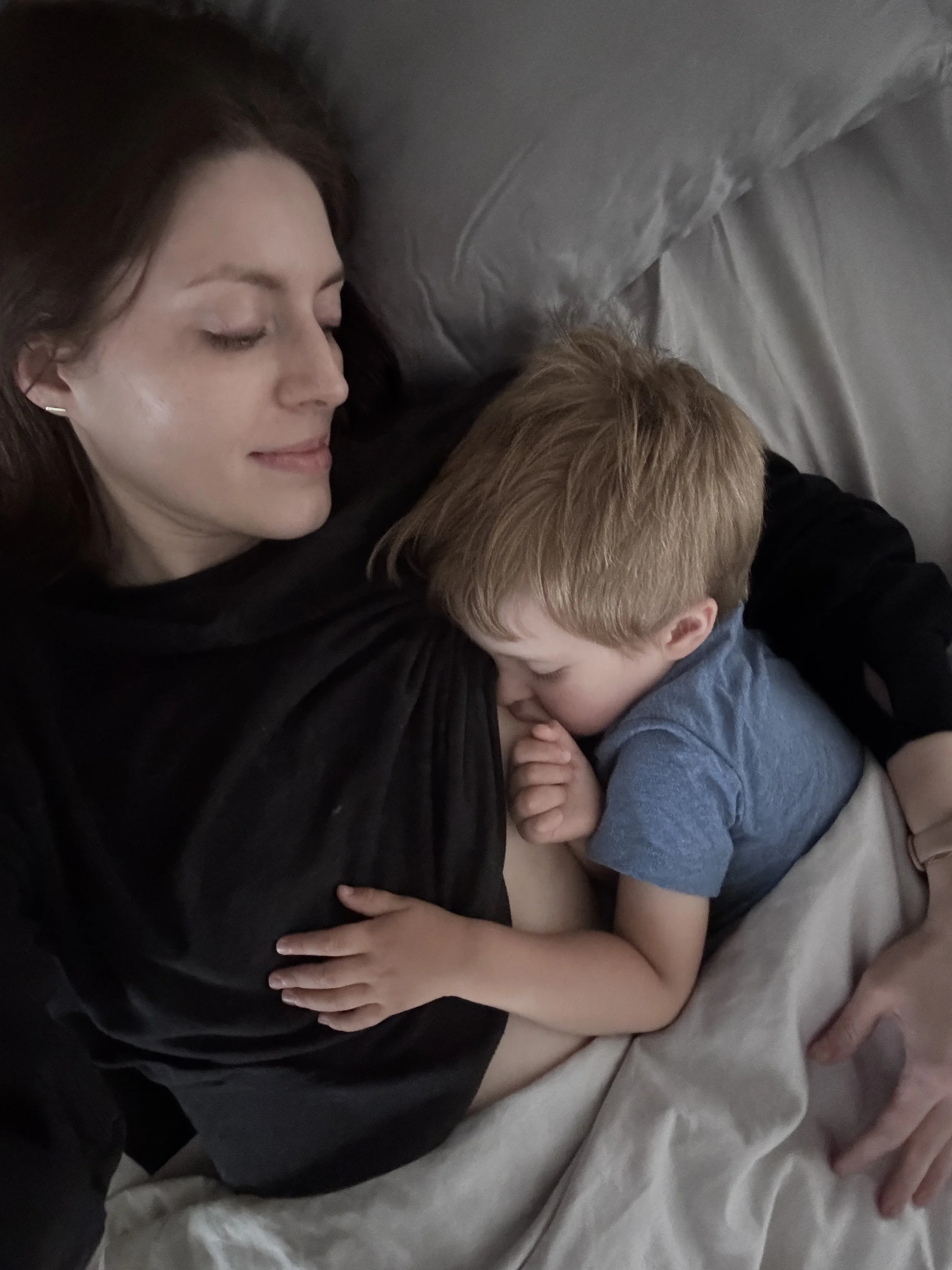 A woman with dark hair lying on a bed with her eyes closed, hugging a young boy with light hair who is resting his head on her chest.