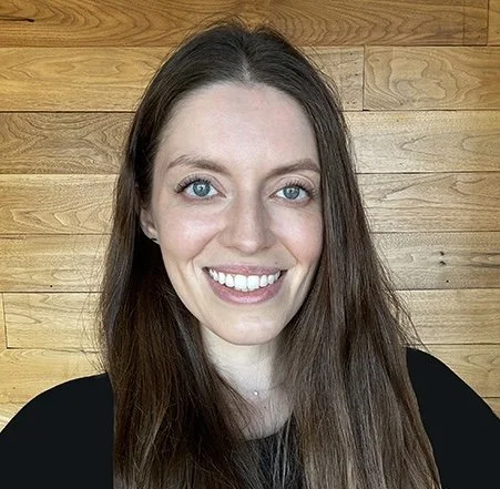 A woman with long brown hair and blue eyes smiling in front of a wooden wall.