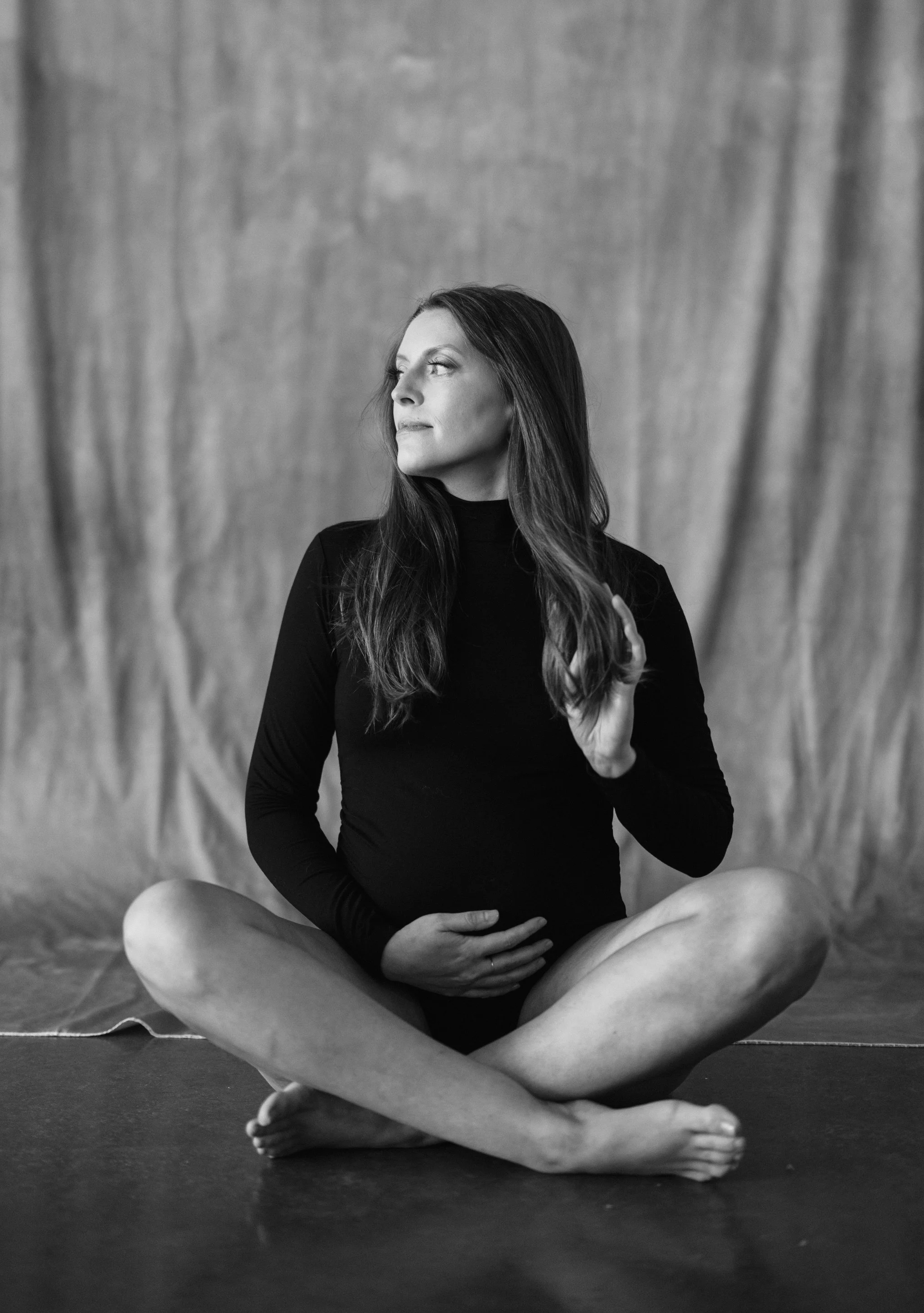 Black and white photo of a woman sitting cross-legged on the floor, wearing a long-sleeved black top, with her left hand placed on her stomach and her right hand touching her chest, looking to her left, with a plain curtain backdrop.