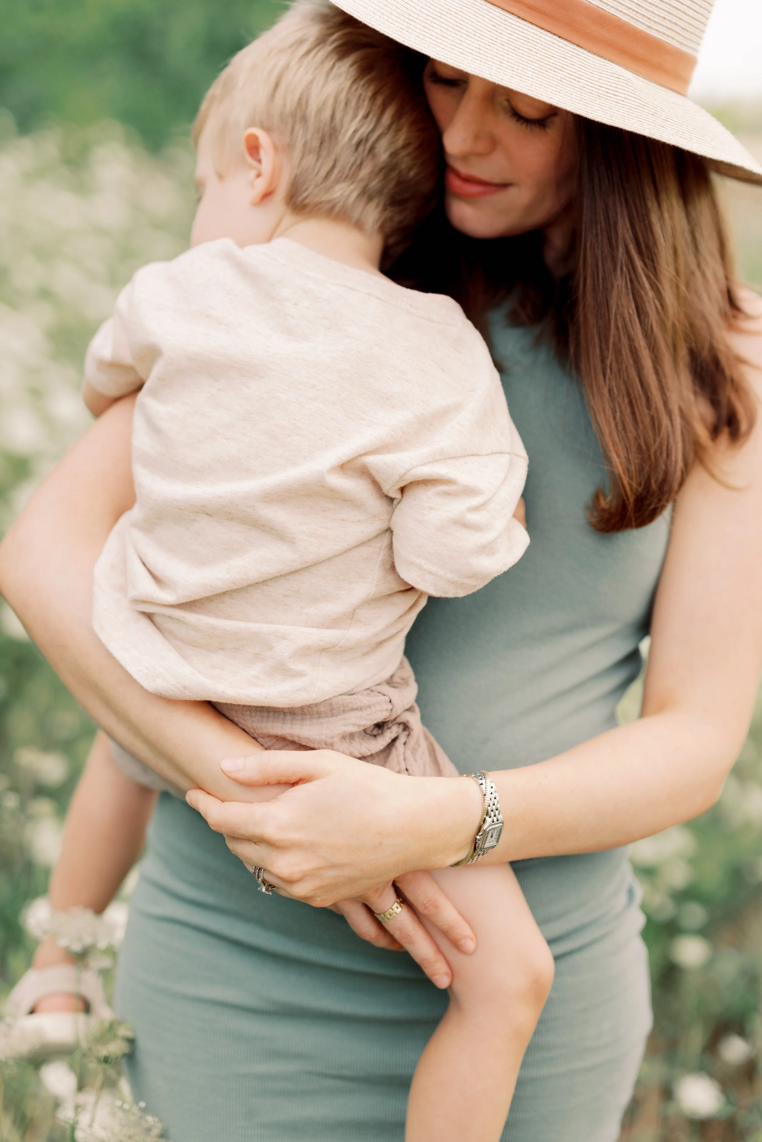 A woman wearing a wide-brimmed hat holding a young boy in a garden or outdoor area with blurred greenery and white flowers in the background.
