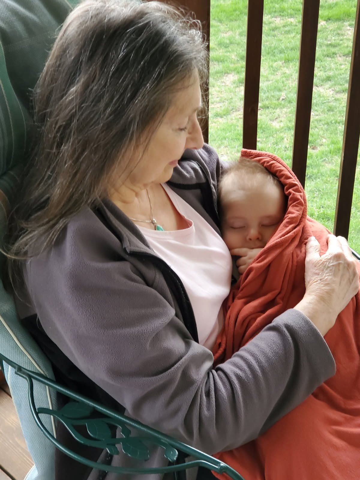 An older woman with gray hair sitting outdoors on a porch holding a young child wrapped in an orange blanket, the woman is wearing a gray jacket and white shirt, and the child appears to be sleeping.