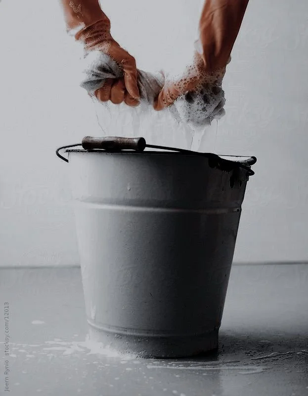 Two hands washing a gray cloth in a metal bucket filled with soapy water.
