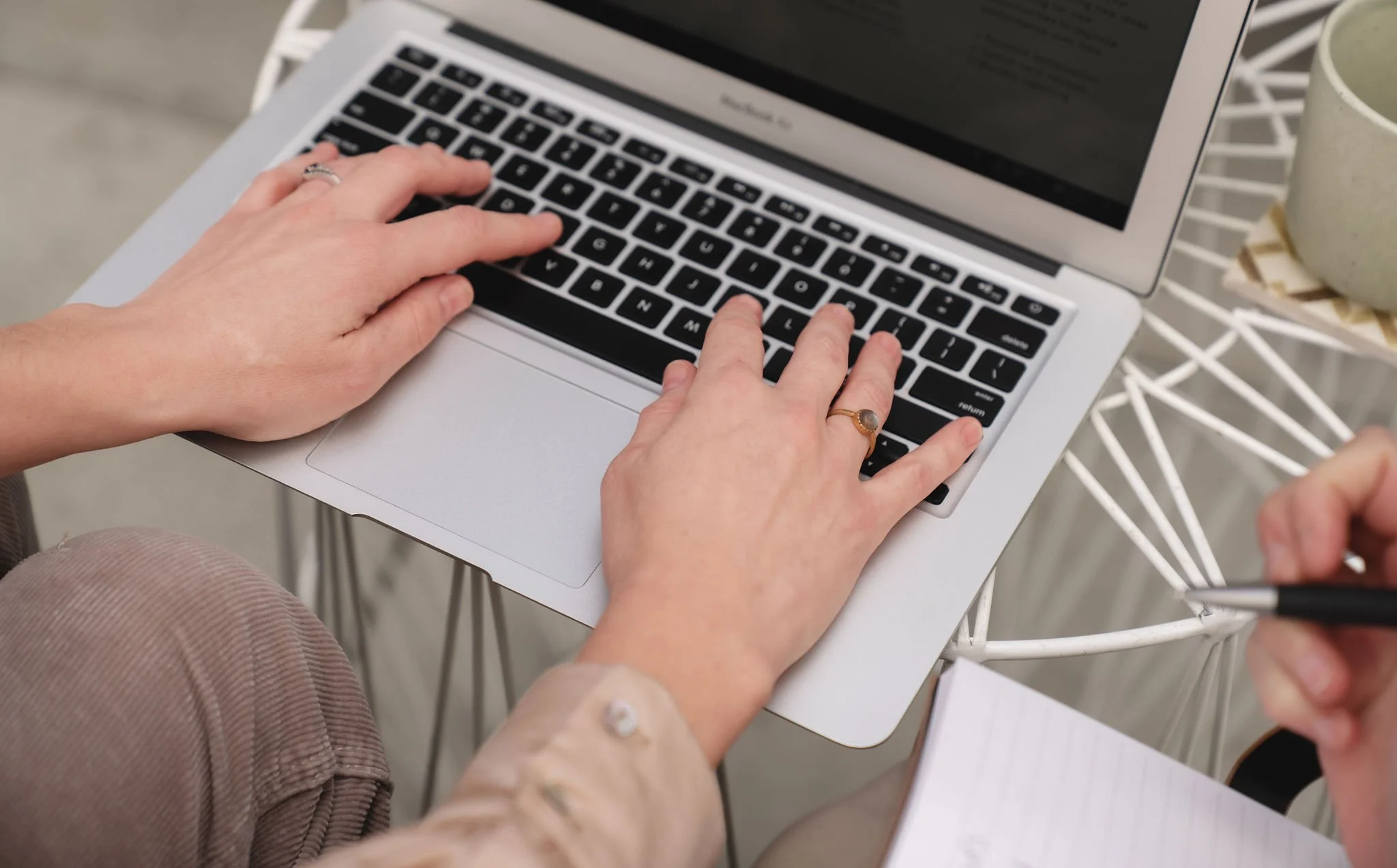 A person typing on a laptop keyboard with both hands, seated at a white wire table, with a mug nearby.