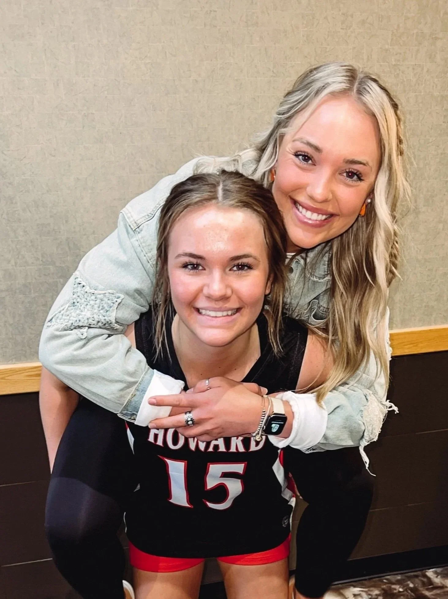 Two women smiling, one wearing a black sports shirt with a number, and the other behind her with blonde hair, a white denim jacket, and orange earrings.