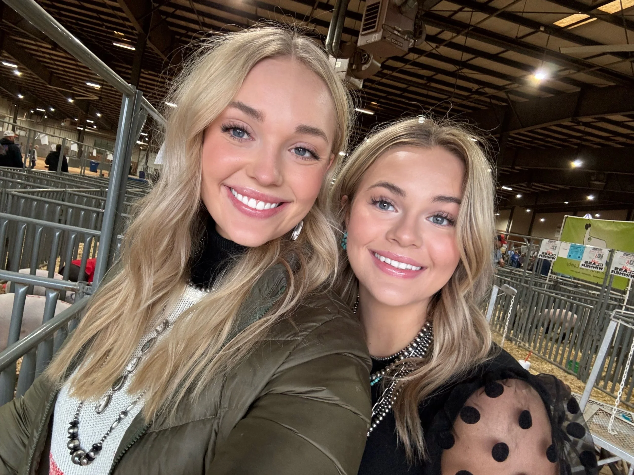 Two women with blonde hair smiling for a selfie in an indoor arena or stadium with metal railings and a high ceiling.