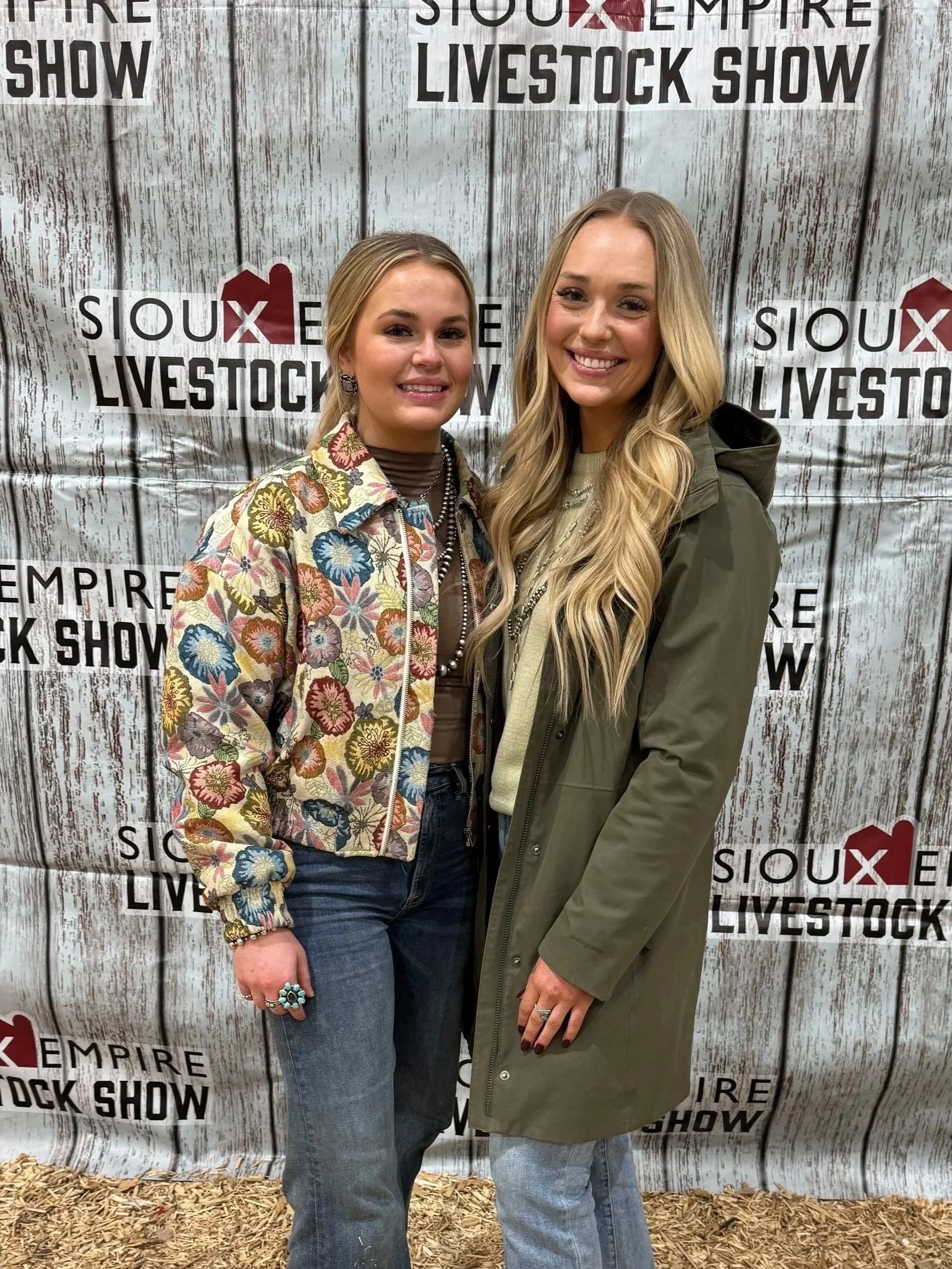 Two women standing in front of a backdrop with the words 'Sioux Empire Livestock Show'. One woman wears a colorful floral jacket and jeans, the other wears a green jacket and jeans. Both are smiling.