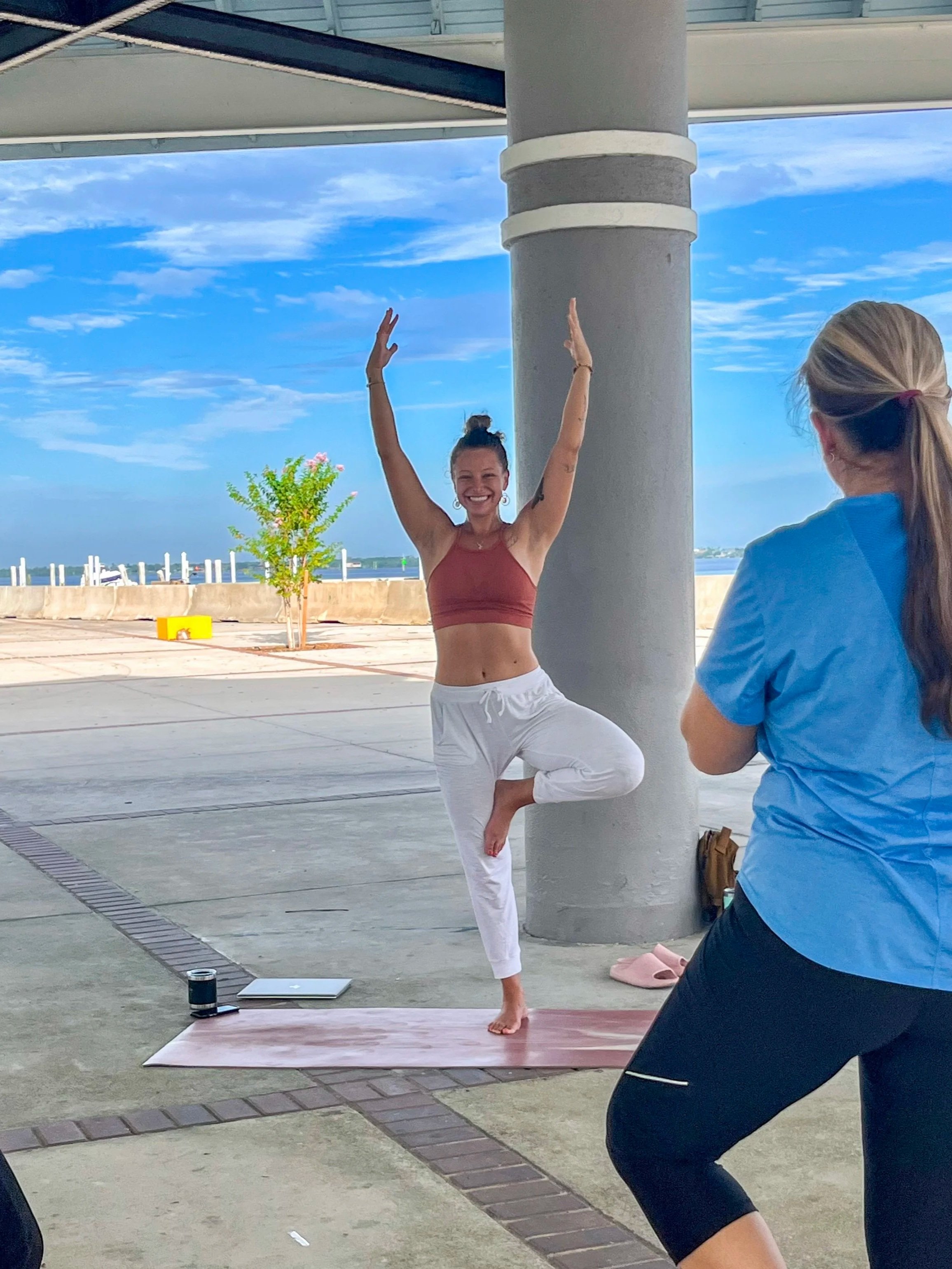 A woman practicing yoga outdoors on a pink mat, wearing a maroon sports bra and white pants, balancing on one leg with her hands raised above her head, while another woman observes in the foreground.