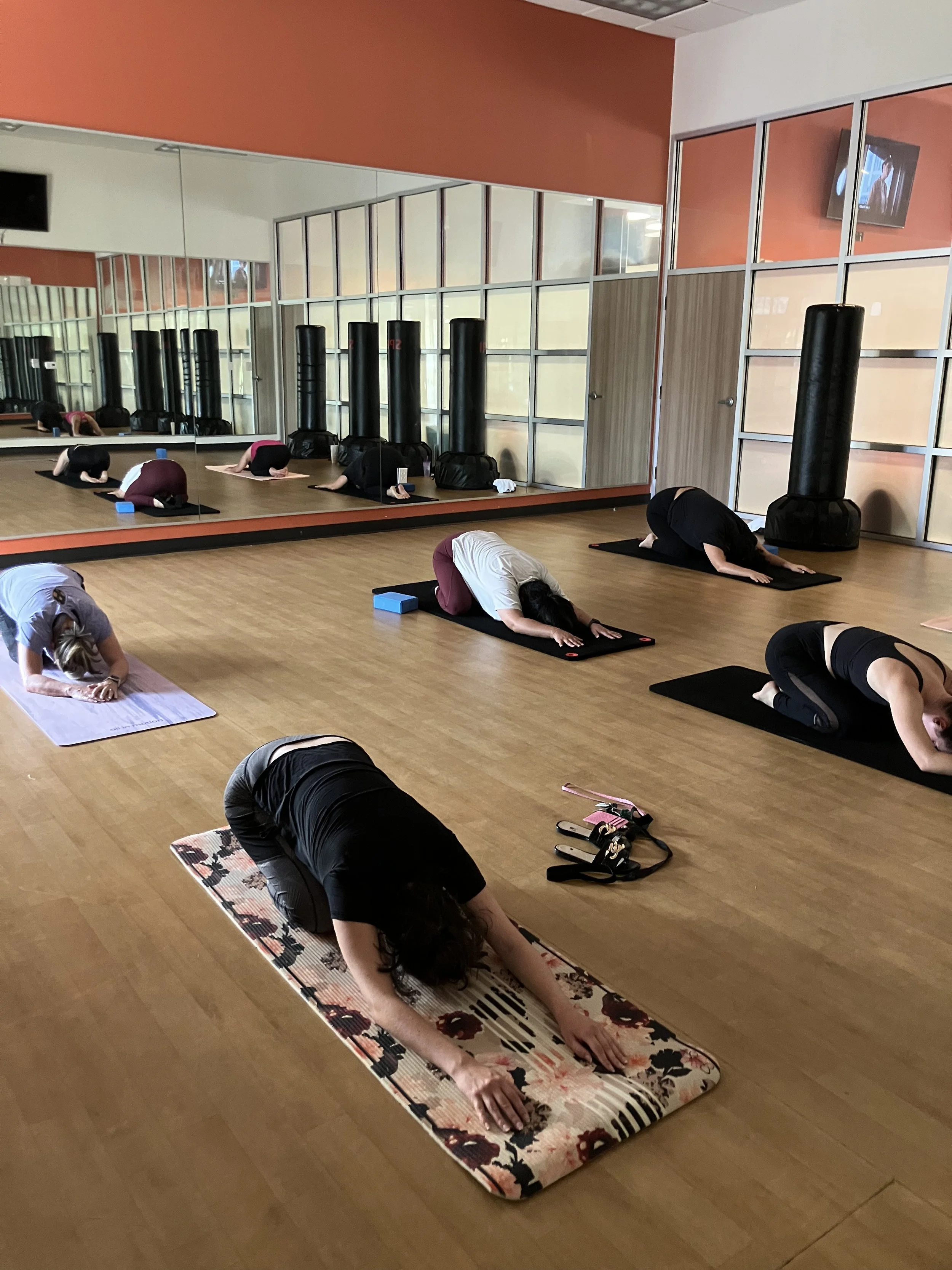 Individuals participating in a yoga class in a fitness studio, performing child poses on yoga mats. The studio has a wooden floor, large mirrors, a wall with a tv, and punching bags.