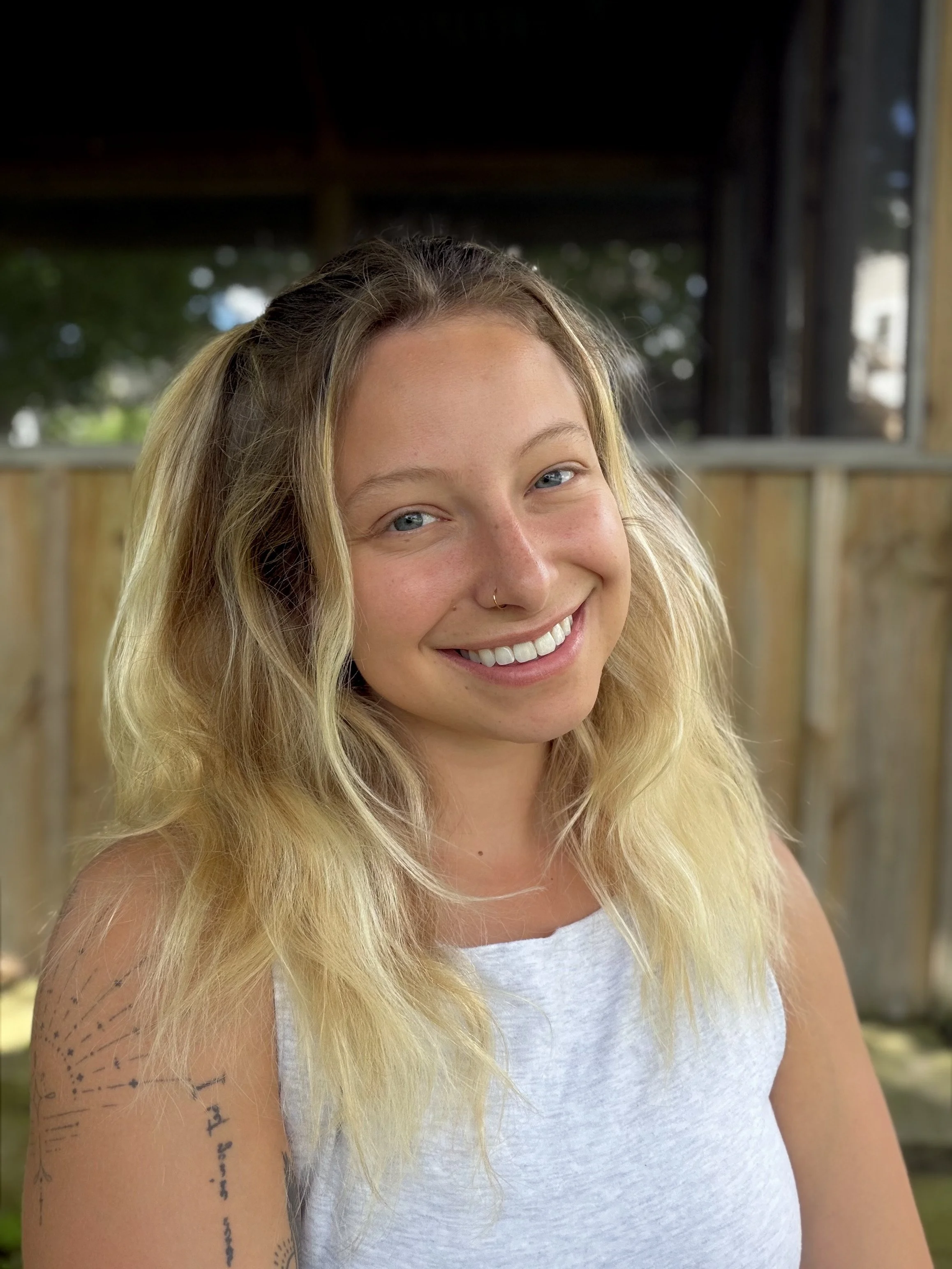 A young woman with blonde hair smiling outdoors, wearing a white tank top, with a tattoo on her arm and a wooden fence in the background.