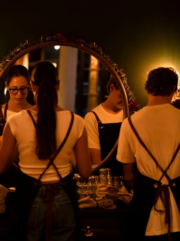 Three people in an indoor setting looking at a mirror, wearing black aprons over white shirts, with glasses and long hair, warm yellow lighting, and a table with glasses and towels in front of them.
