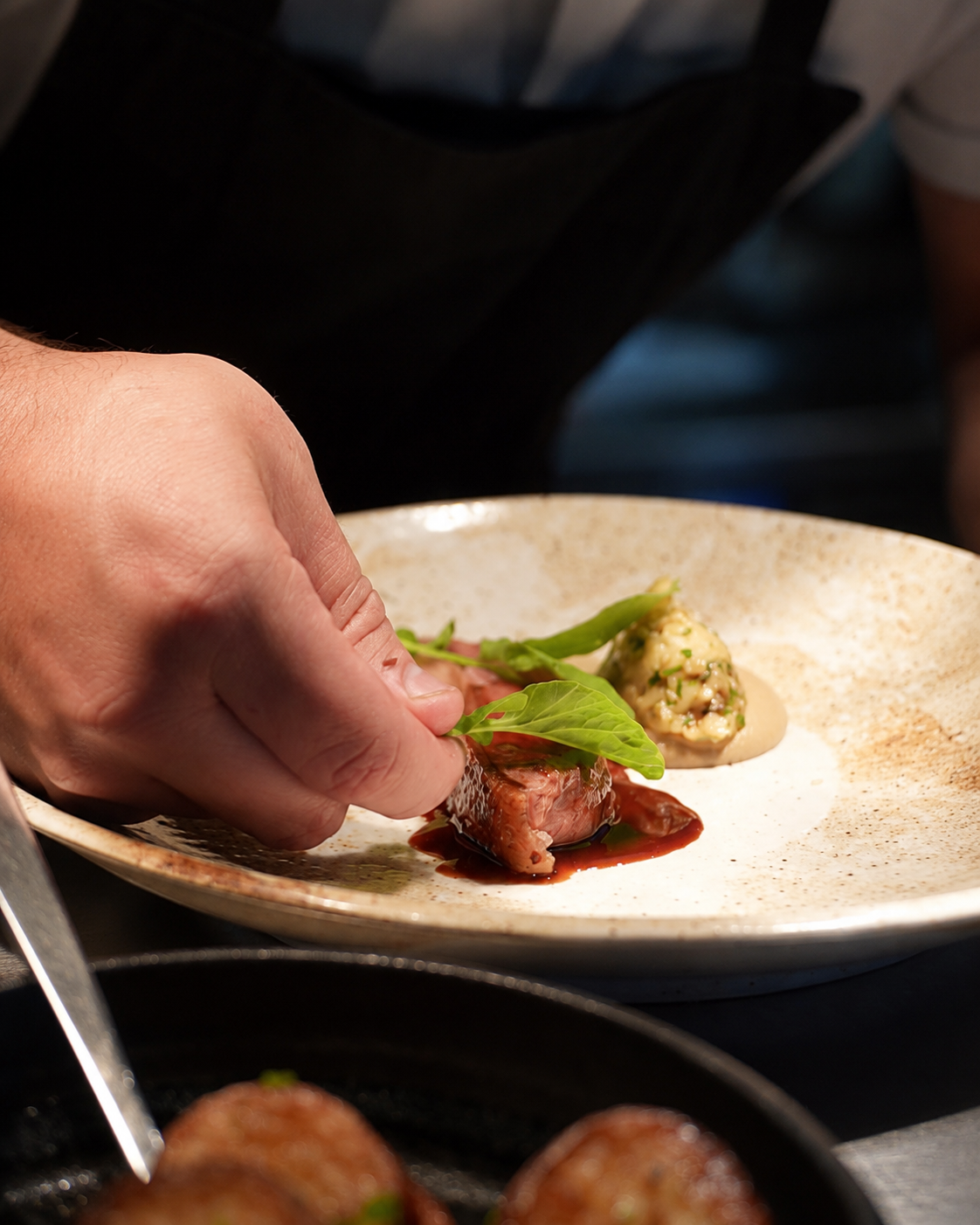 A chef garnishing a plated dish with fresh herbs and a ball of gourmet mousse or terrine, with another dish in the foreground.