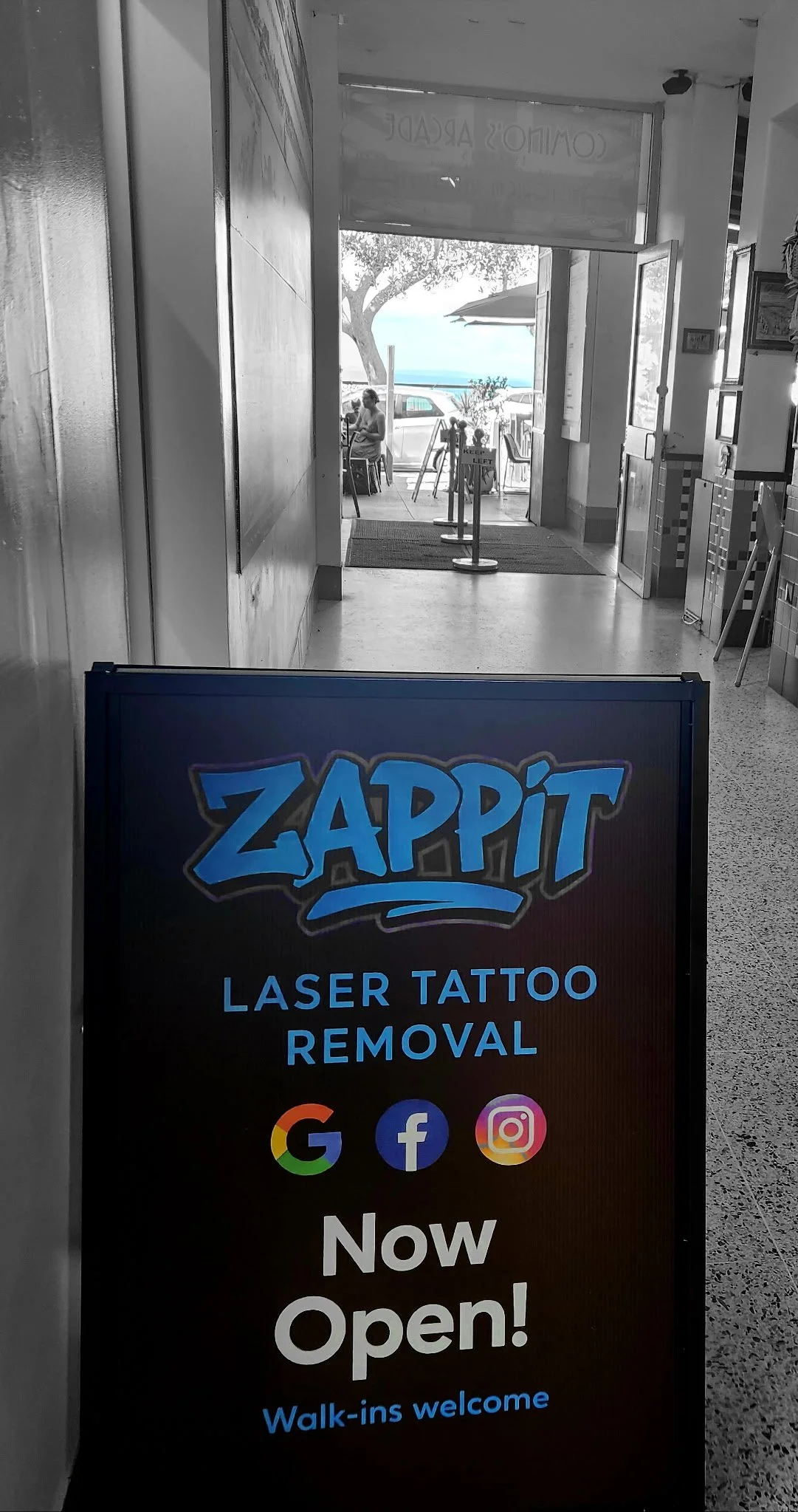 Signboard for Zappit laser tattoo removal shop outside a store, with icons for Google, Facebook, and Instagram, and the message 'Now Open! Walk-ins welcome.' In the background, an open door reveals a seating area with people near the beach, trees, an