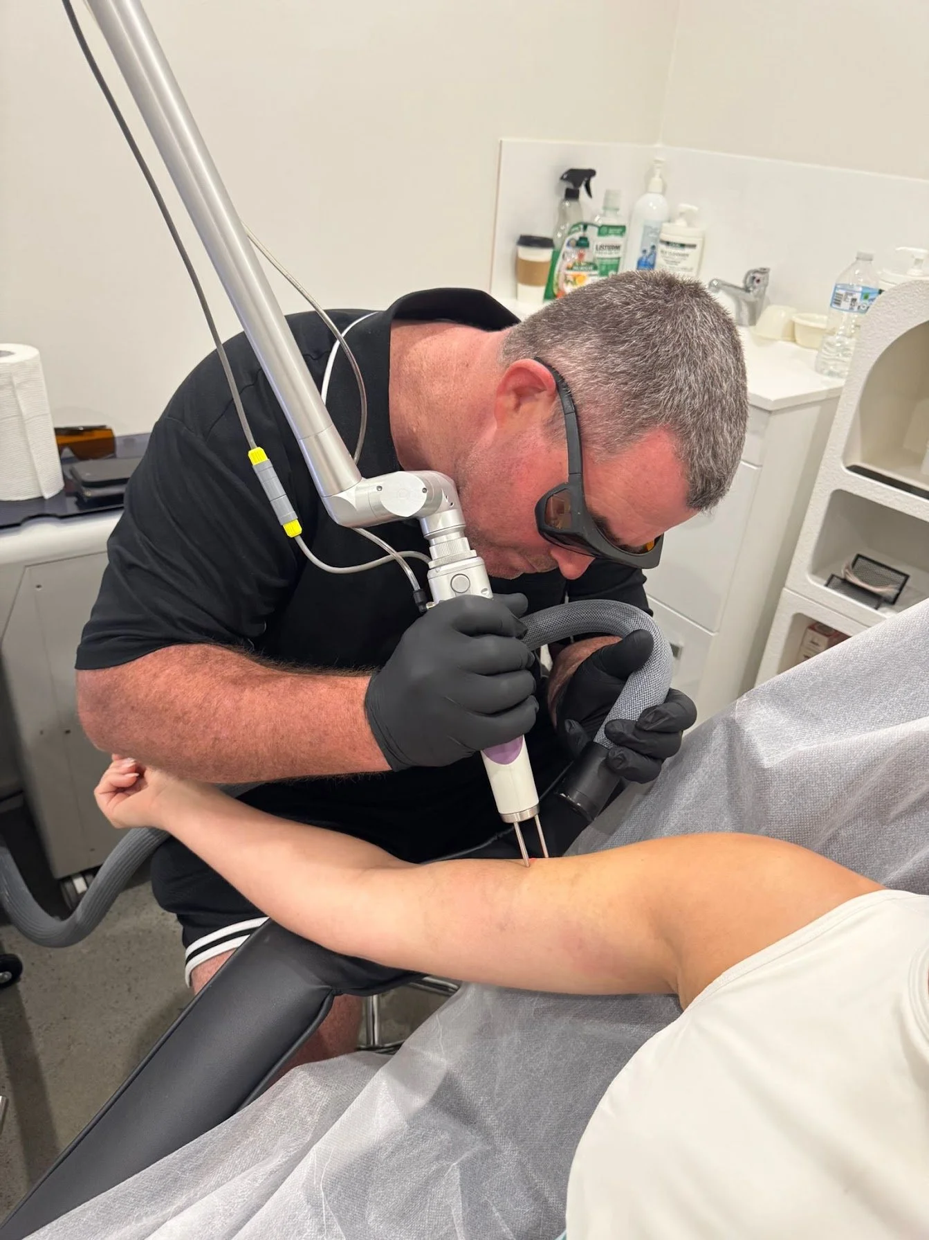 A man with short gray hair, wearing glasses and black gloves, is performing a medical procedure using a specialized device on a patient's arm in a clinical setting.