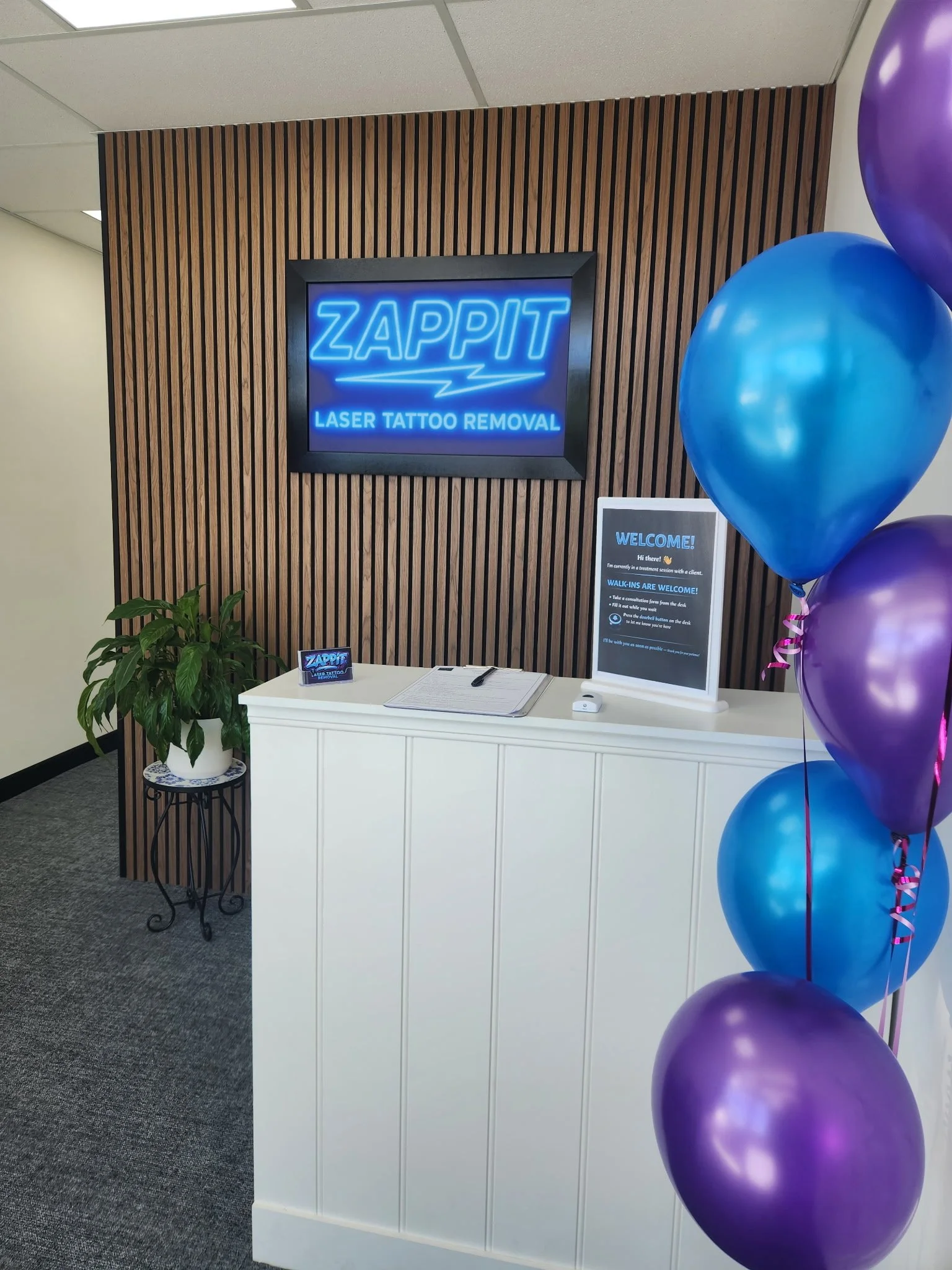 Reception desk at Zappit Laser Tattoo Removal clinic with neon sign, welcome sign, potted plant, and blue and purple balloons