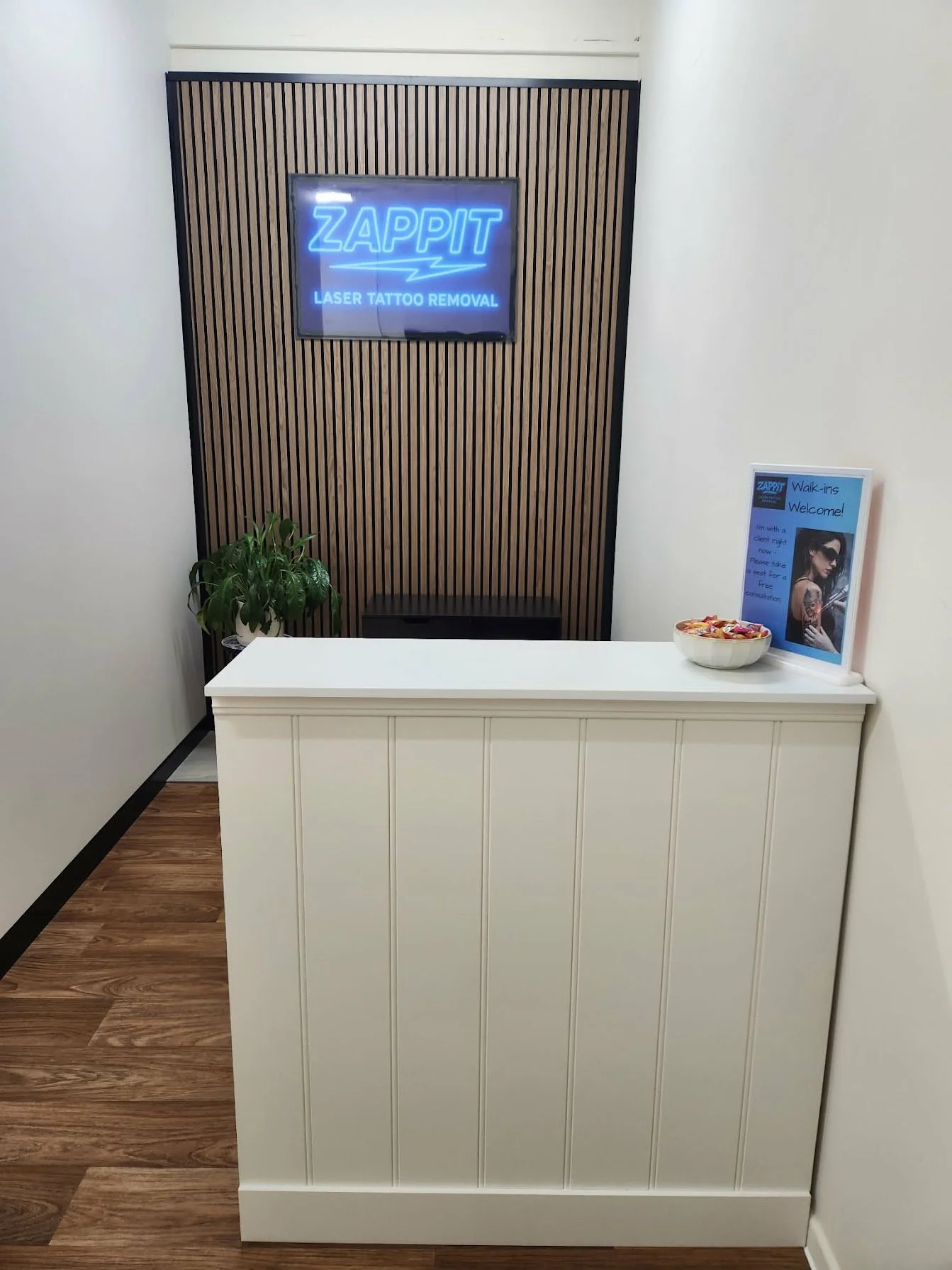 Reception area with a white counter, a potted plant, a bowl of candies, and a sign welcoming walk-ins for laser tattoo removal with a woman holding a tattoo gun.