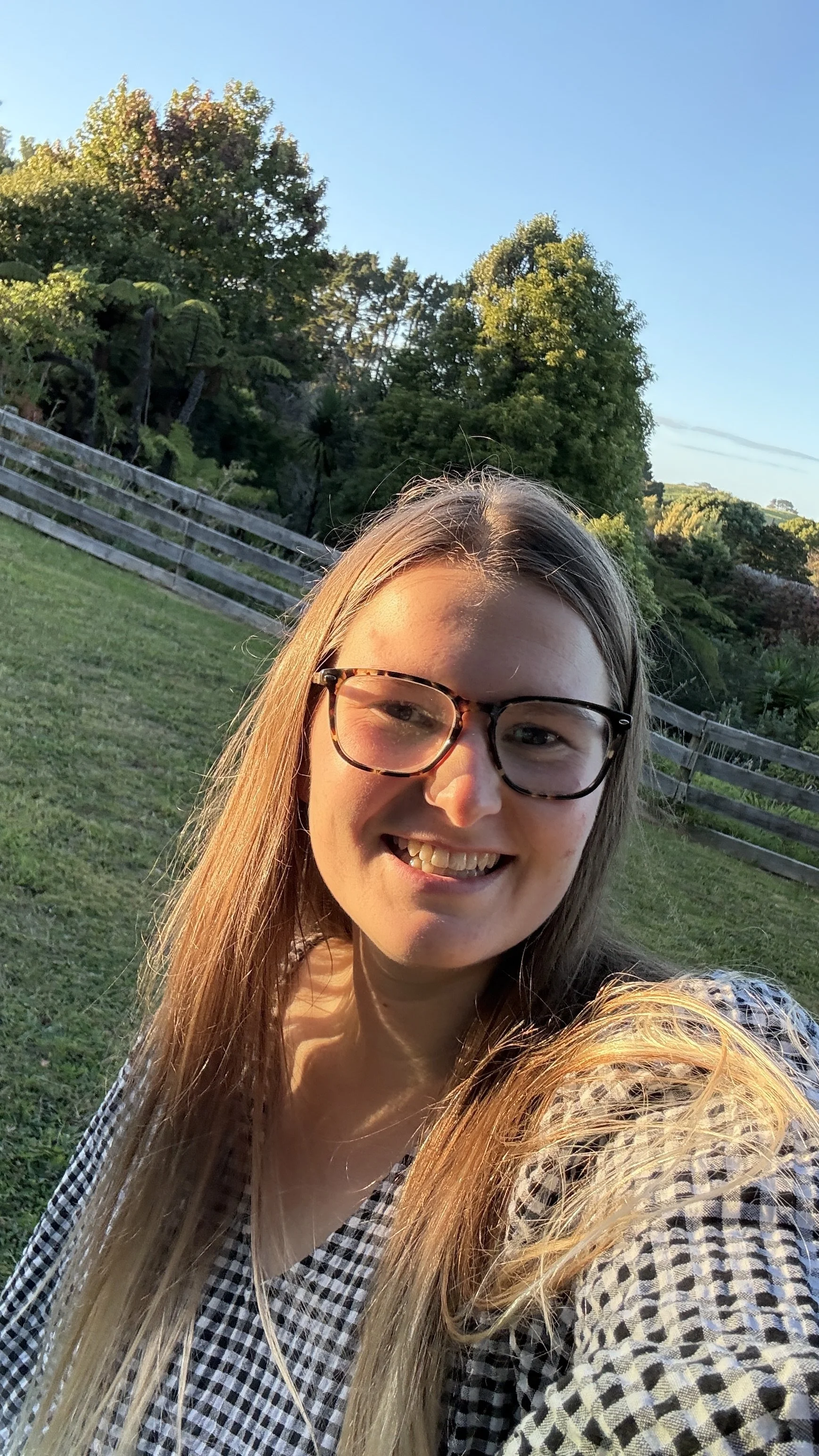 Brooke Druyven, smiling woman with long blonde hair and glasses, outdoors with green trees, wooden fence, and blue sky in the background.