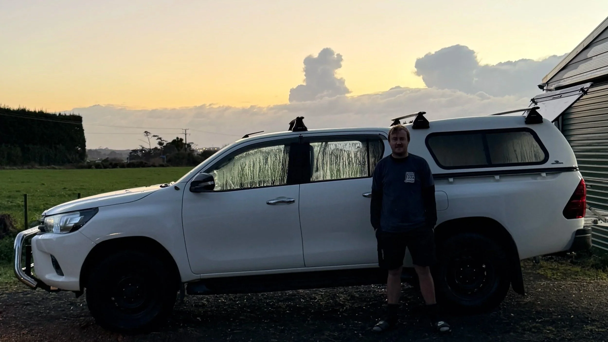 Reece Ewing, owner of Ewing Plumbing, standing next to a white SUV with a canopy, in a rural setting during sunset, with cloudy sky in the background.