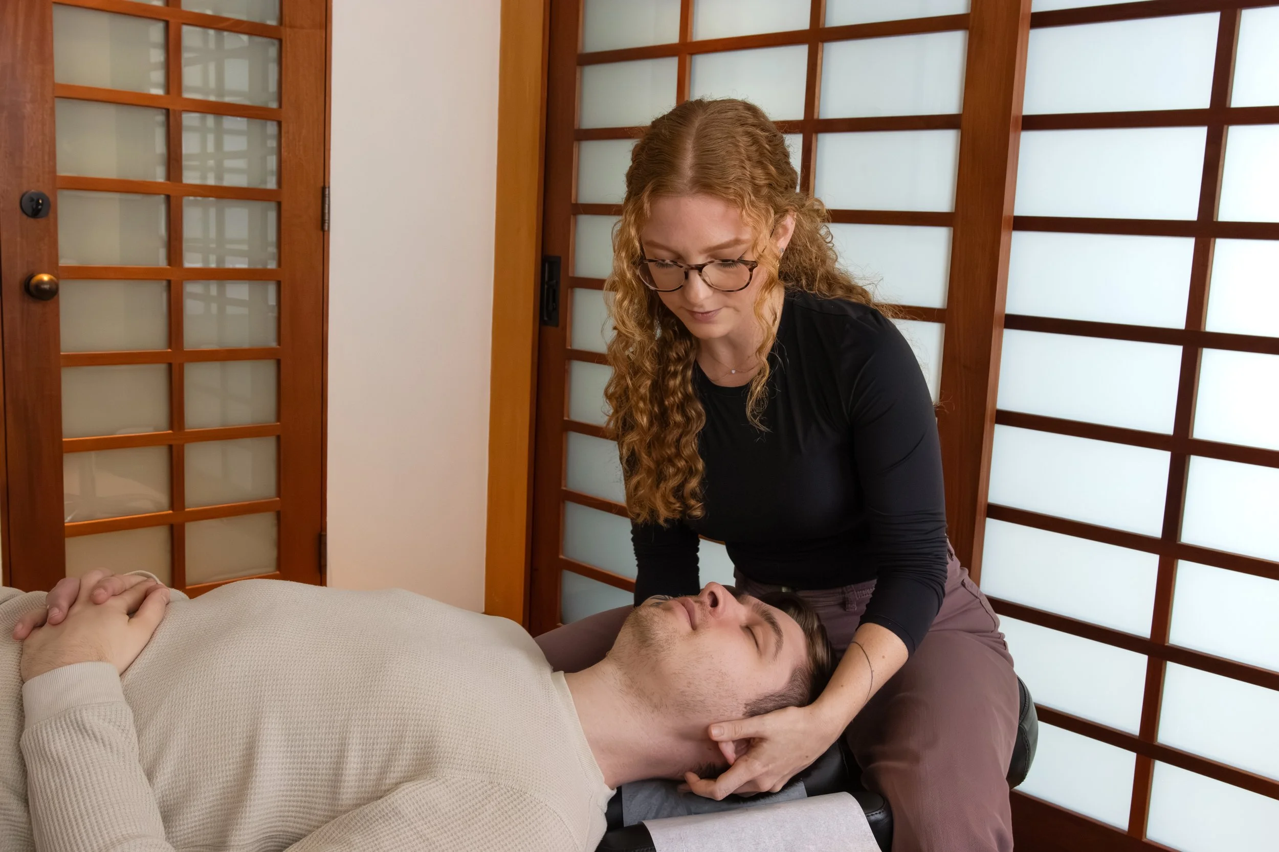 A woman with glasses and curly red hair performing reiki healing on a man lying peacefully with his eyes closed in a room with wooden sliding doors.