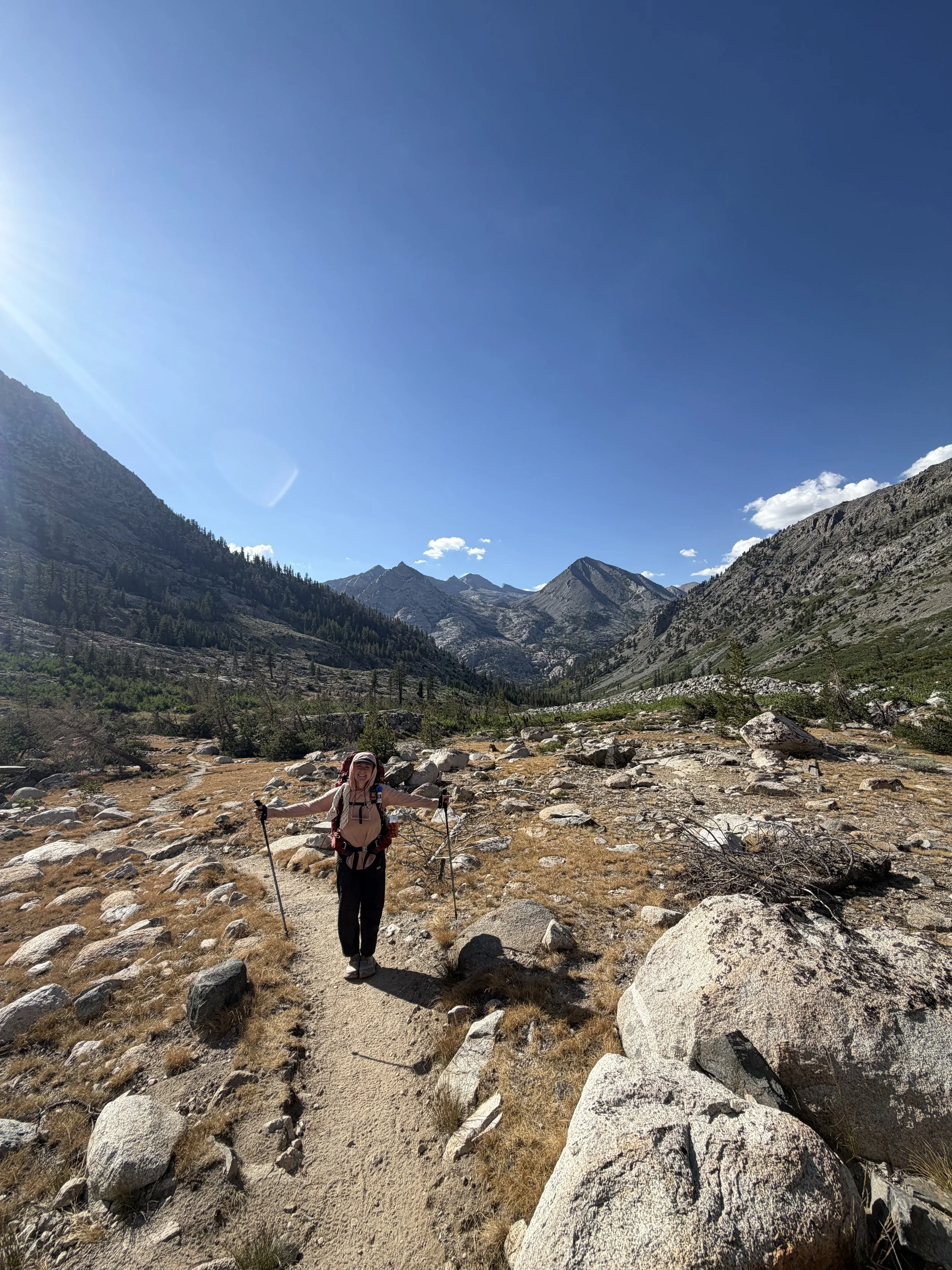 A woman hiking in a mountainous area with rocky terrain, wearing outdoor gear and holding hiking poles, surrounded by mountains under a clear blue sky. Trail running, hiking, backpackinathlete, sports chiropractic, rehab for outdoor athletes.