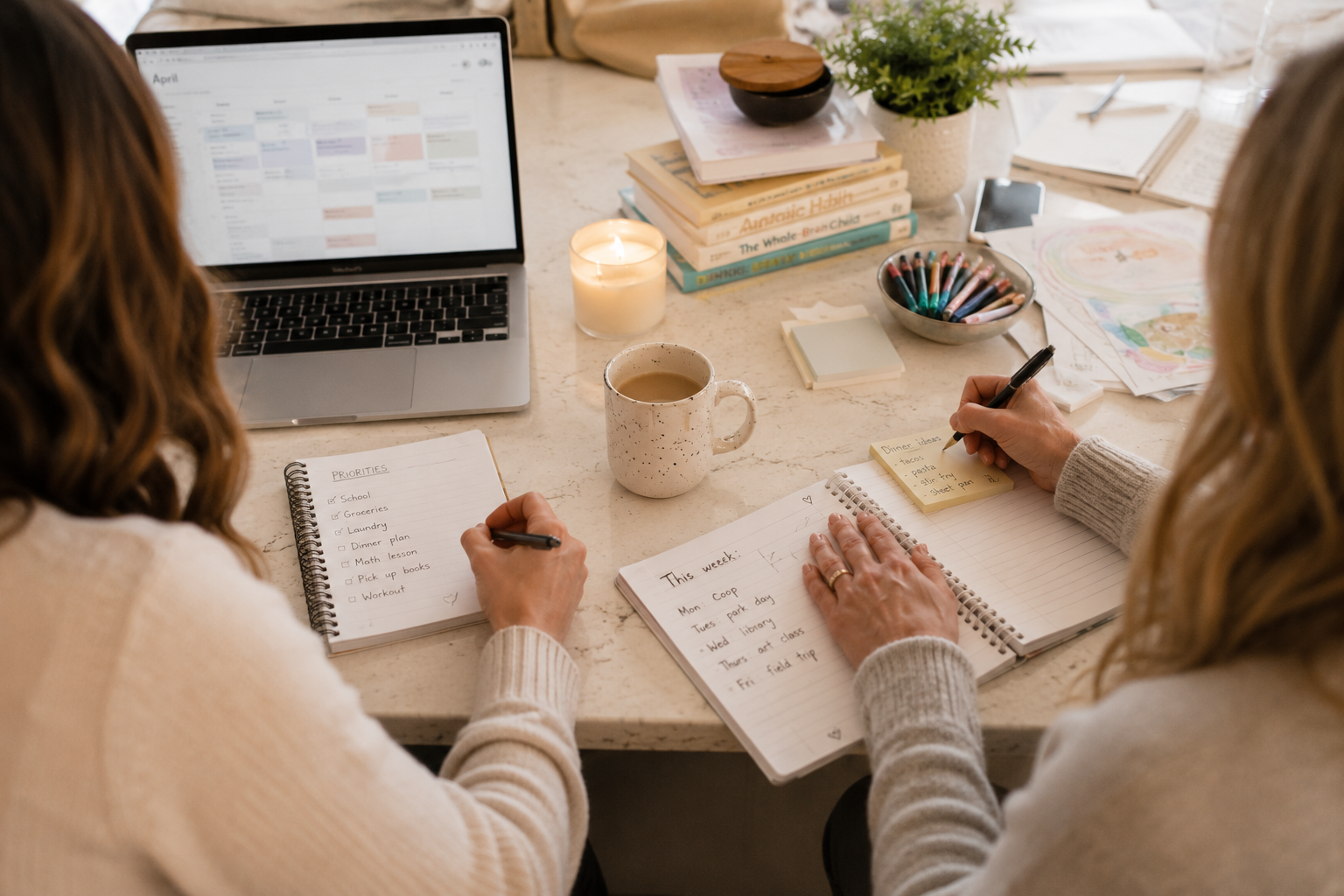 Two women sitting at a table working on planners and notebooks with a laptop, books, a candle, a mug, colored pens, and artwork on the table.