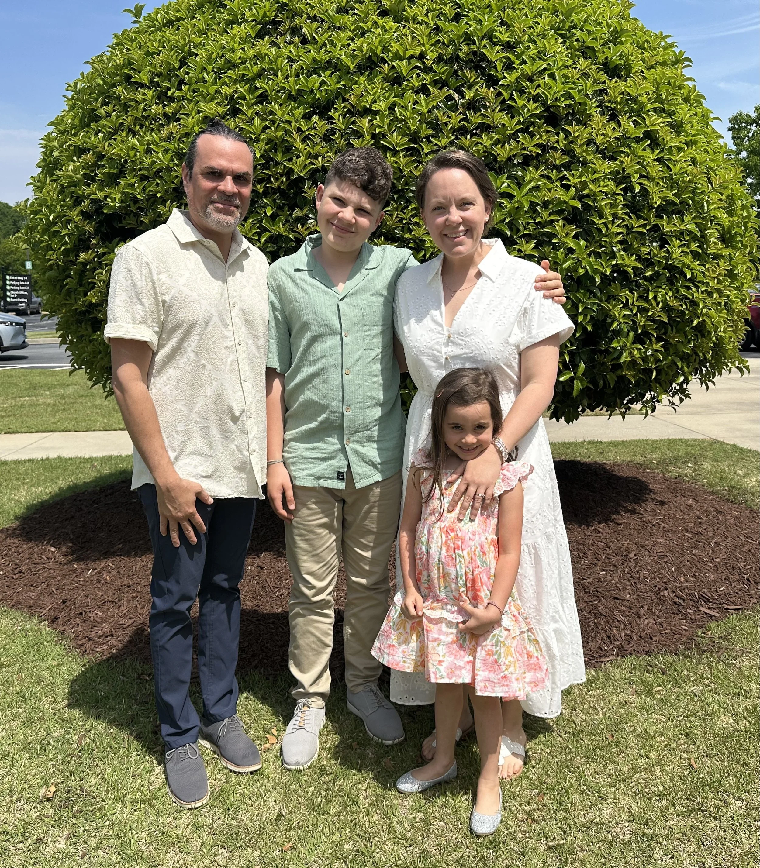 Family of five standing outdoors on grass in front of a large green bush, smiling. The family includes two men, two women, and two young girls.