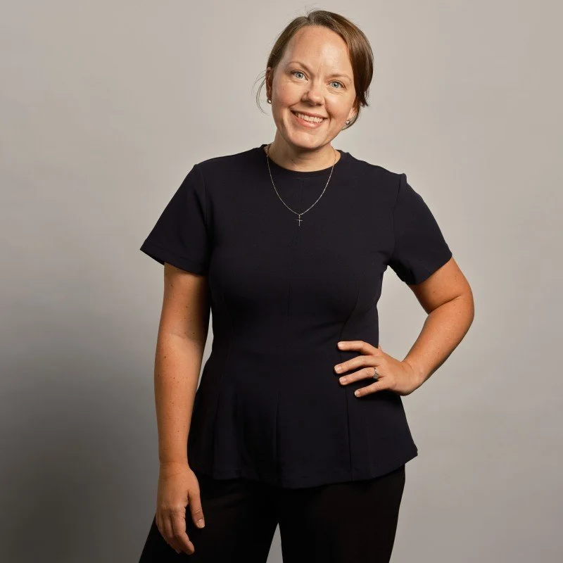 Smiling woman with short brown hair, wearing a black dress and a necklace with a cross pendant, standing with one hand on her hip against a neutral gray background.