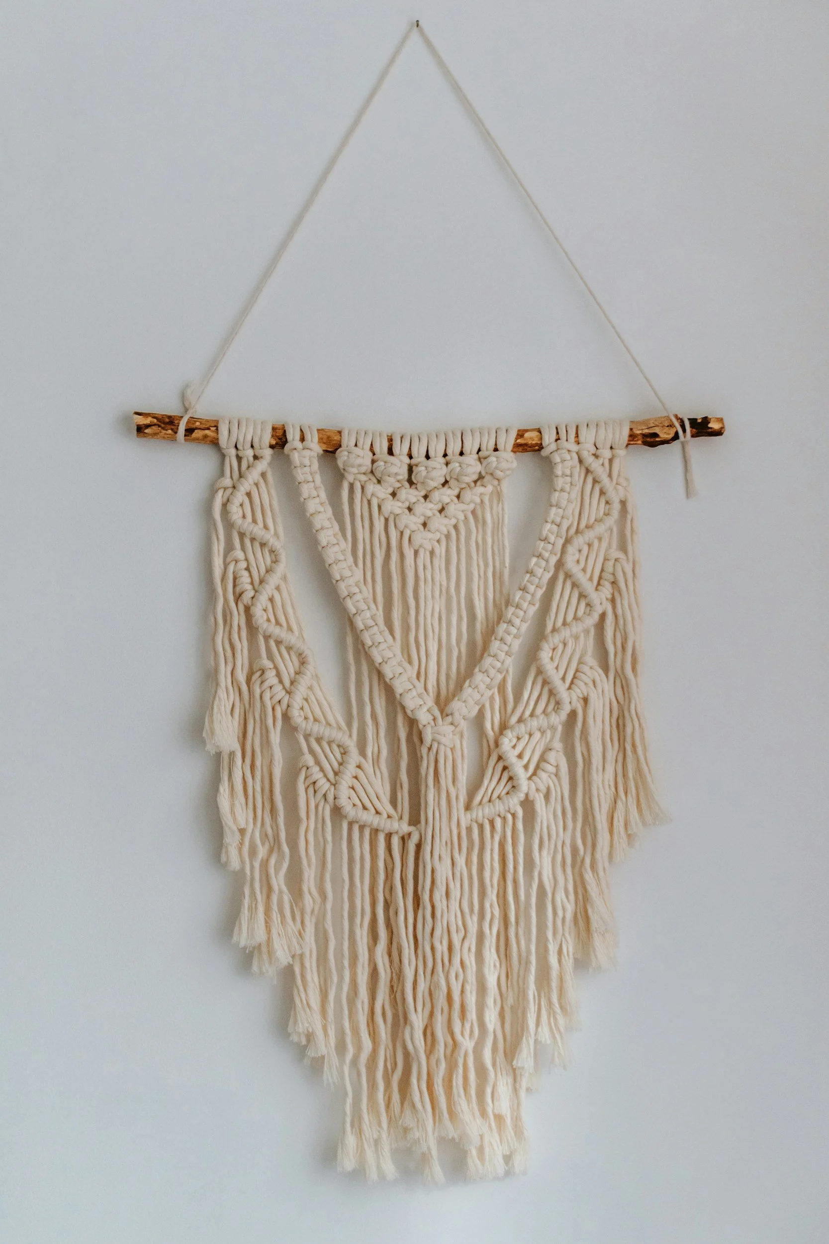 A wall hanging made of a wooden stick and cream-colored yarn, featuring intricate macrame patterns and tassels. It is suspended from a string attached to a hook on a plain white wall.