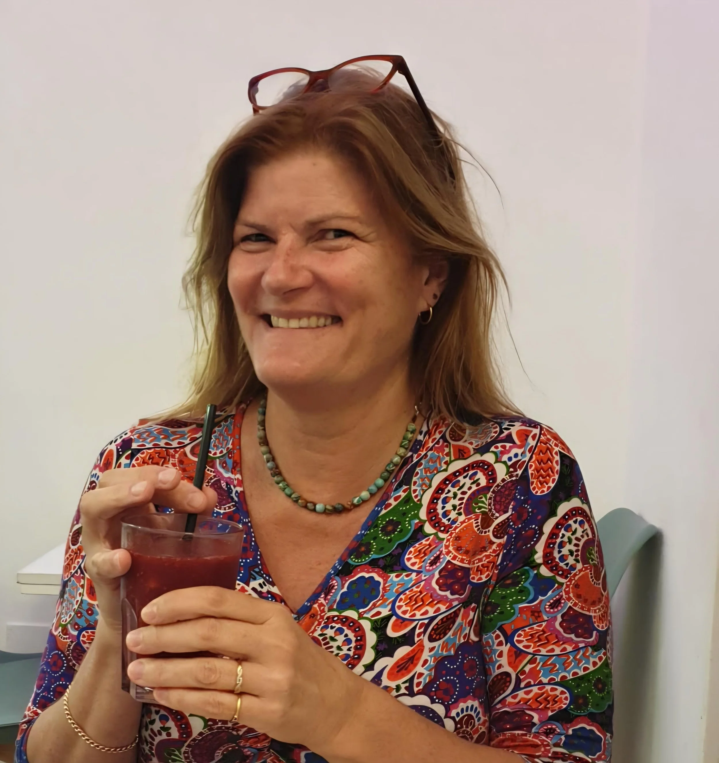A woman with red hair, glasses on her head, and multiple jewelry pieces, smiling while holding a glass of red drink with a straw, sitting in a room with a white wall and a chair.