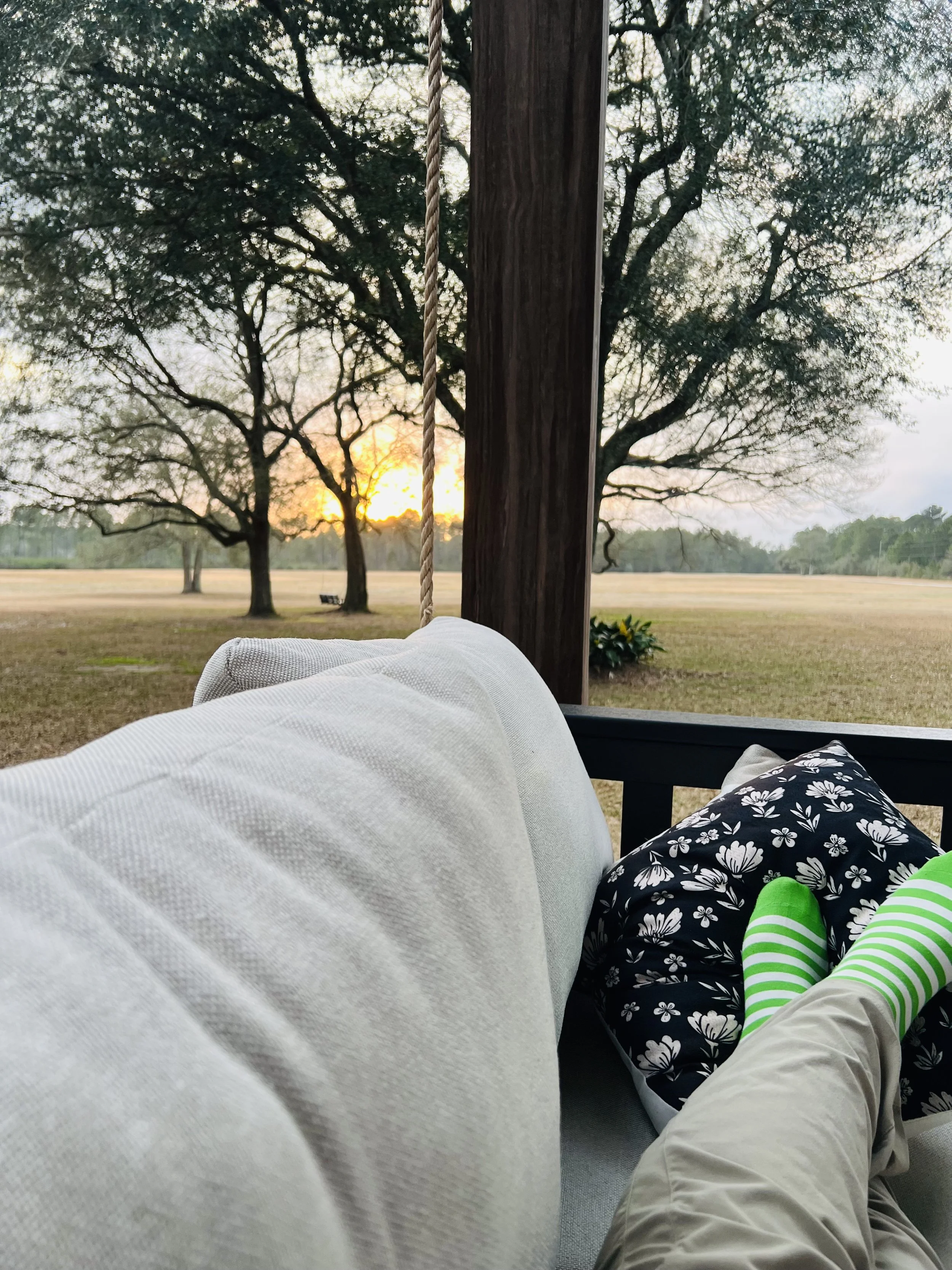 Person relaxing on a porch swing, looking out at a scenic view of trees and a sunset in the distance.