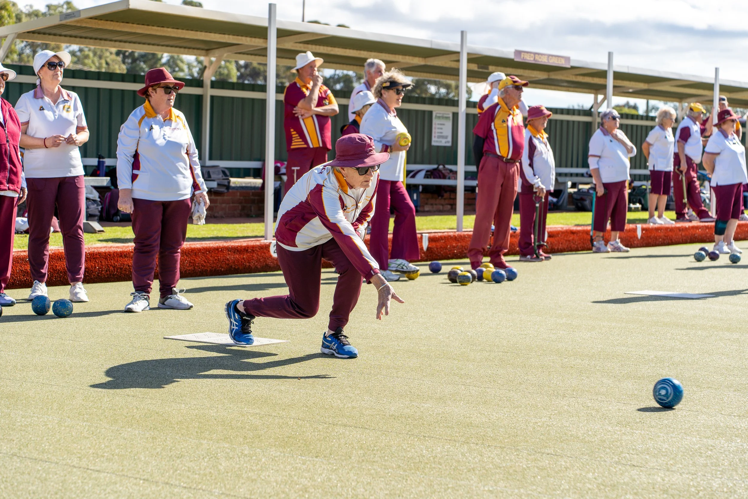 Group of seniors playing lawn bowls outdoors on a sunny day, with some preparing to throw bowls and others watching.