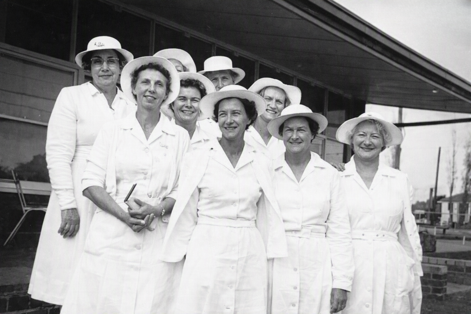 Black and white photo of nine women wearing white dresses and wide-brimmed hats outdoors.