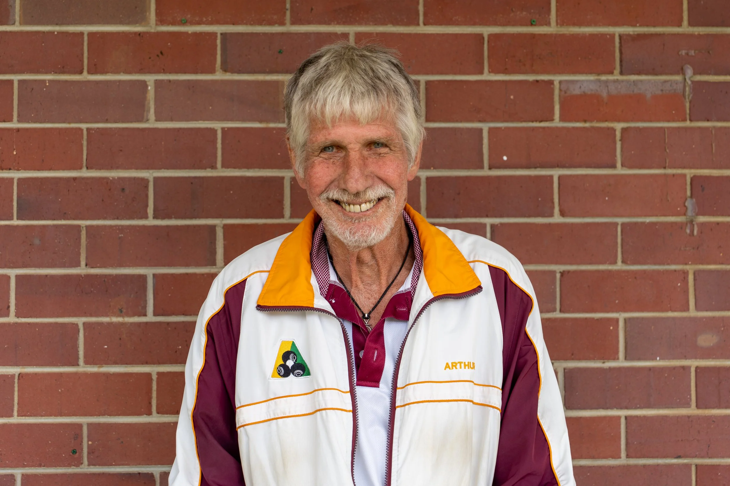 Smiling elderly man with gray hair and beard standing in front of a brick wall, wearing a maroon and white jacket with yellow accents and patches.