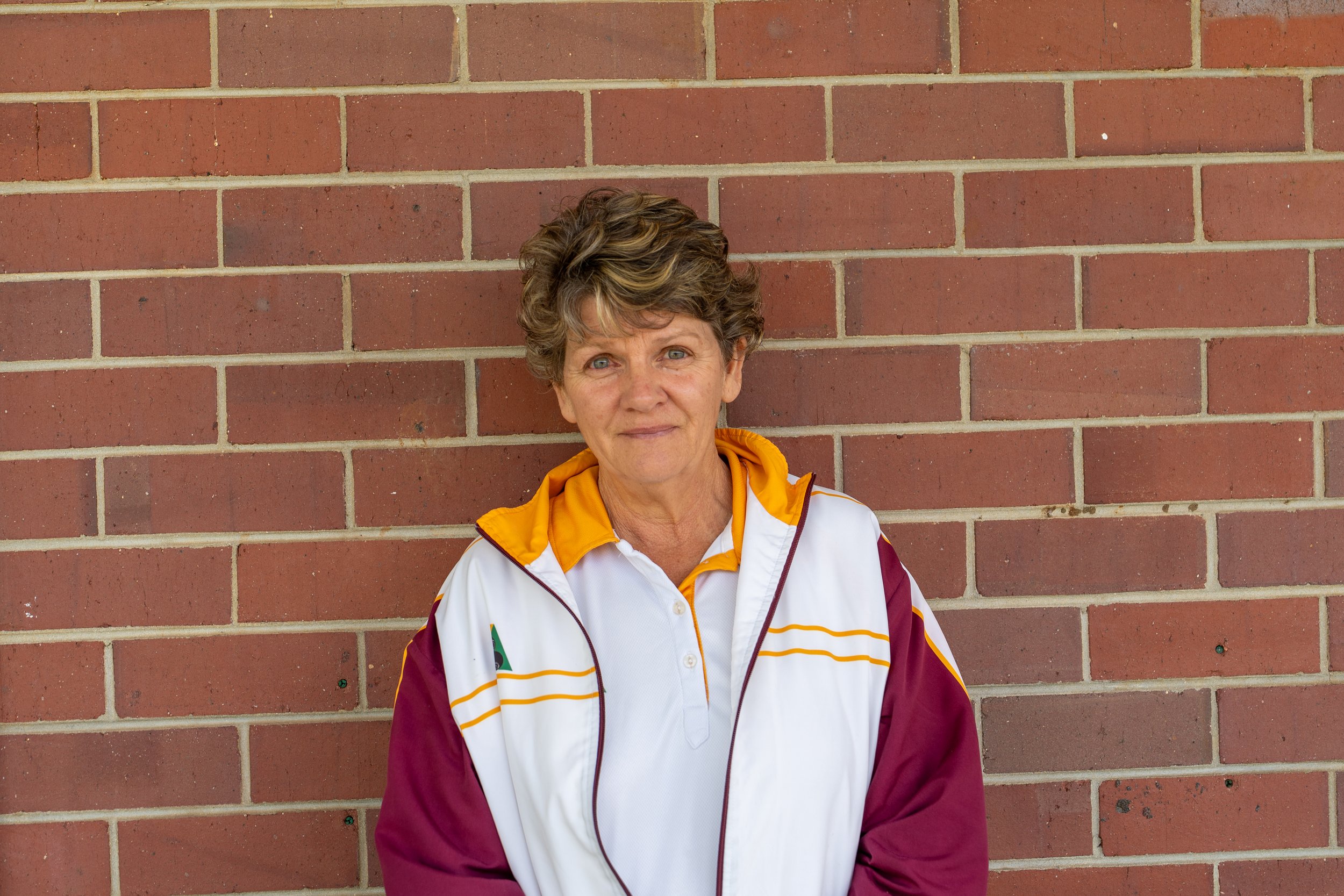 An older woman with short, curly brown hair and blue eyes standing in front of a red brick wall. She is wearing a colorful sports jacket with maroon, white, and yellow accents.