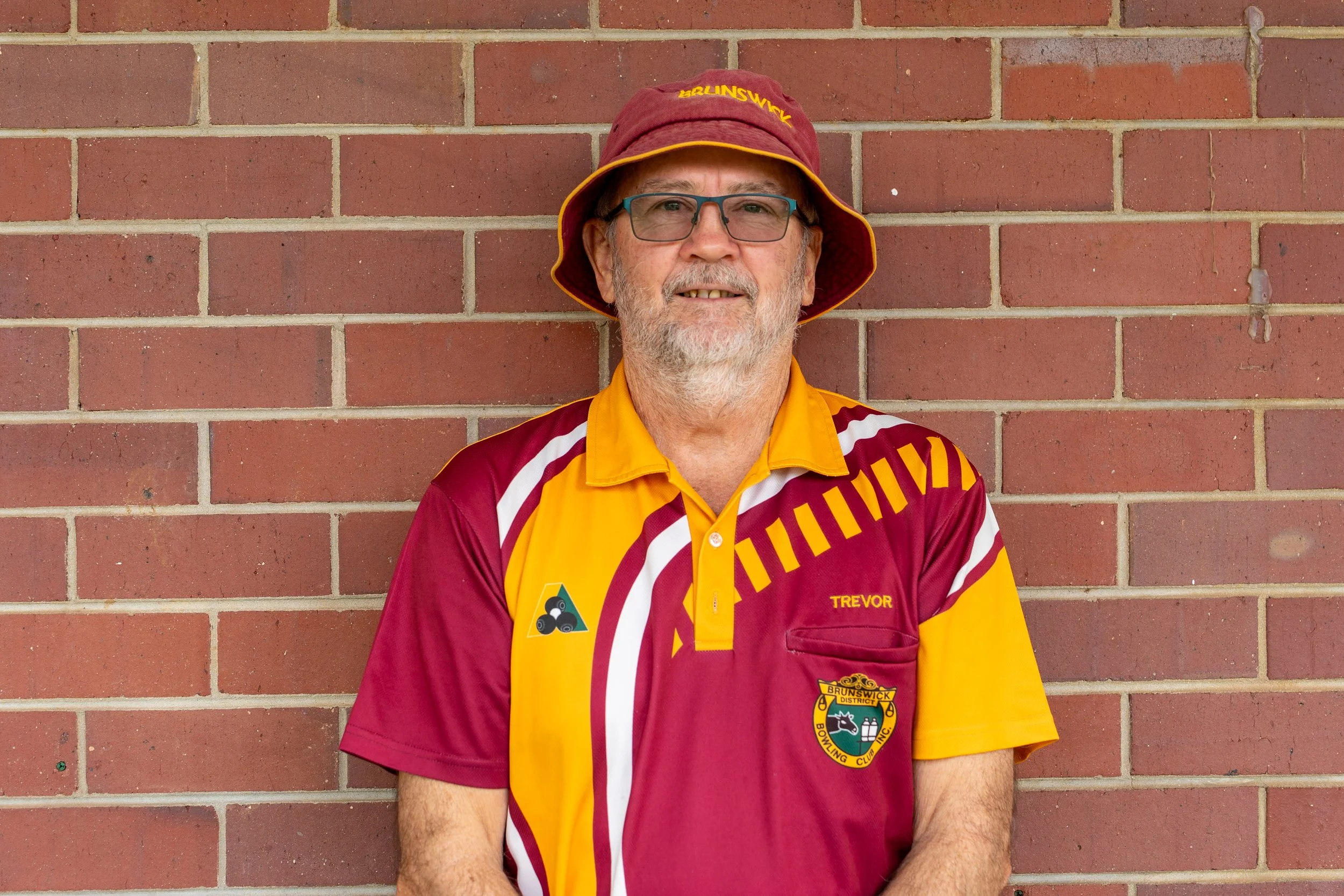 A man with gray hair, glasses, and a beard, wearing a maroon and yellow sports shirt and a maroon bucket hat with yellow lettering, standing in front of a brick wall.