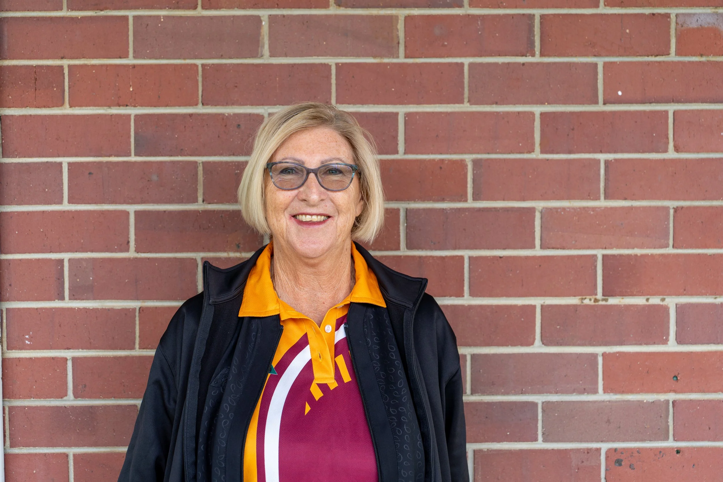 A smiling woman with blonde hair wearing glasses, a maroon and yellow shirt, and a black jacket standing in front of a brick wall.