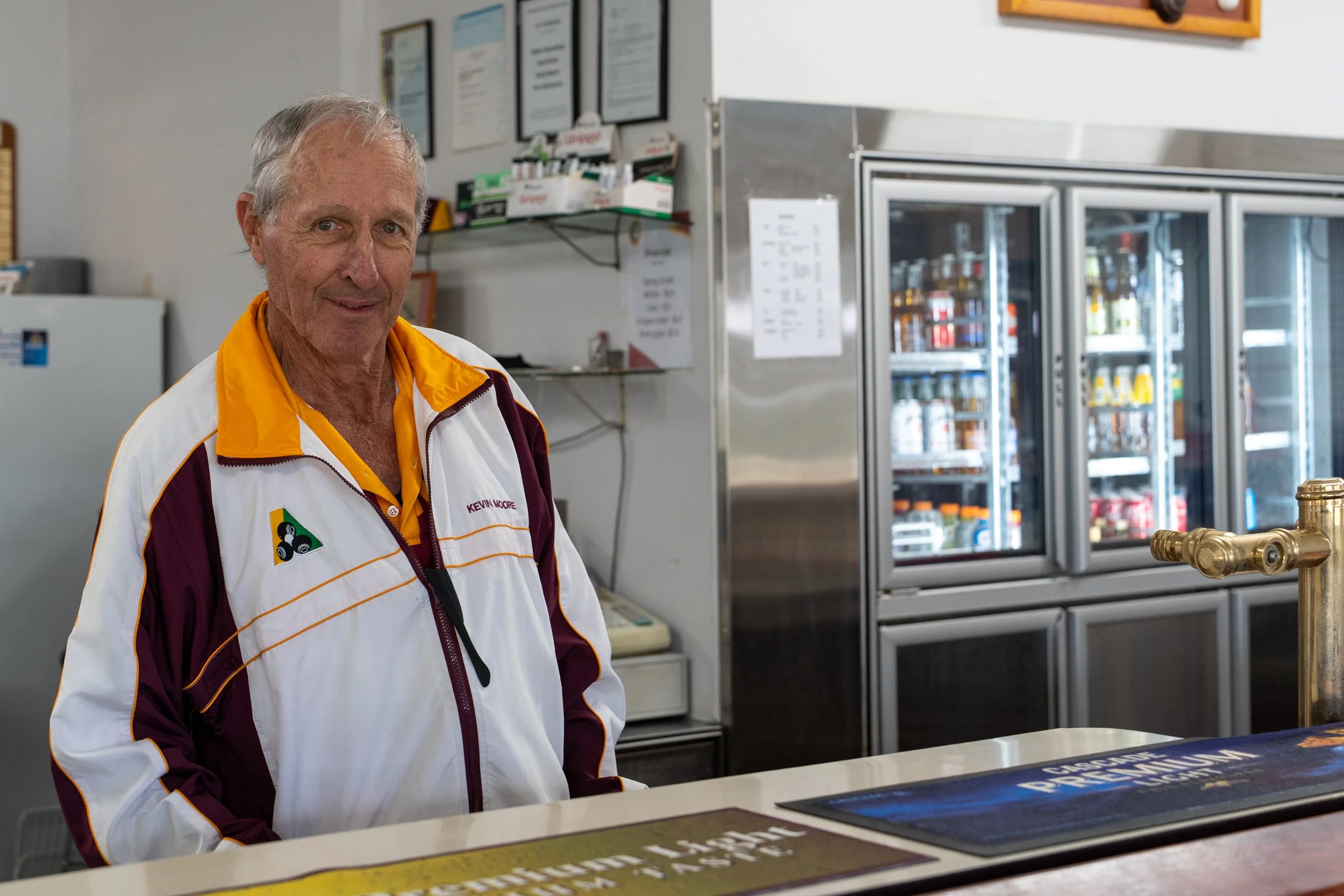 An older man with gray hair, wearing a white and maroon jacket with yellow accents, stands behind a counter in a store or café. In the background, there are framed certificates or notices on the wall and a refrigerator filled with refrigerated beverages.