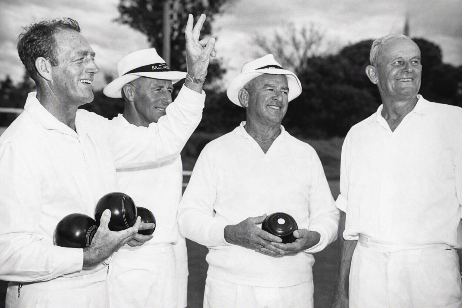 Four elderly men in white clothing and hats holding bowling balls, standing outdoors, with trees in the background, smiling and looking cheerful.