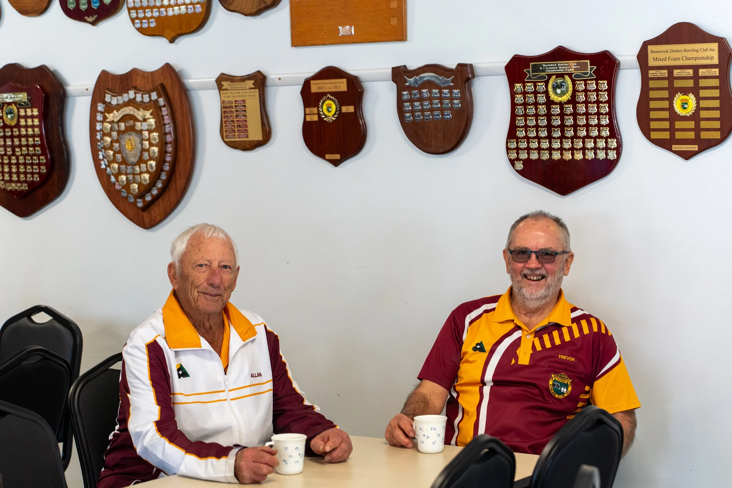 Two smiling men wearing team jerseys sit at a table with cups in hand, in front of a wall decorated with numerous plaques and awards.