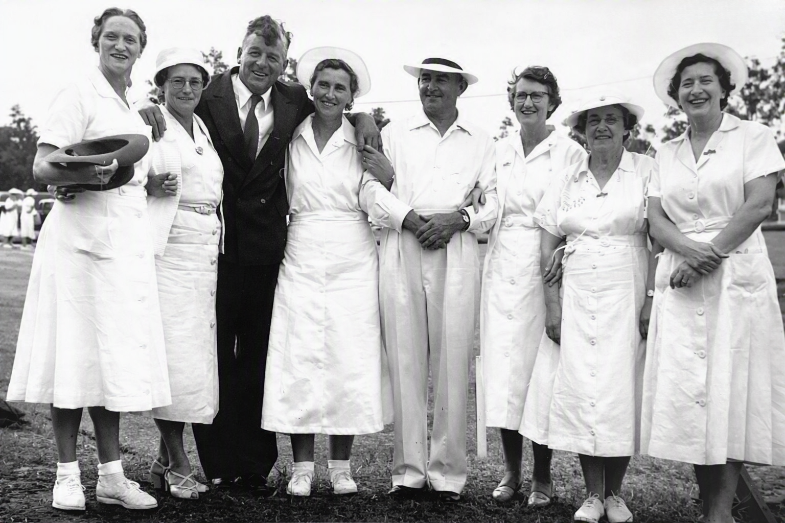 A black and white photo of eight women and two men standing outdoors, dressed in vintage clothing. The women are wearing white dresses and hats, while the men are in suits and hats. They are smiling and appear to be part of a group or team, with some of them holding hats. The background shows a group of people and some trees.