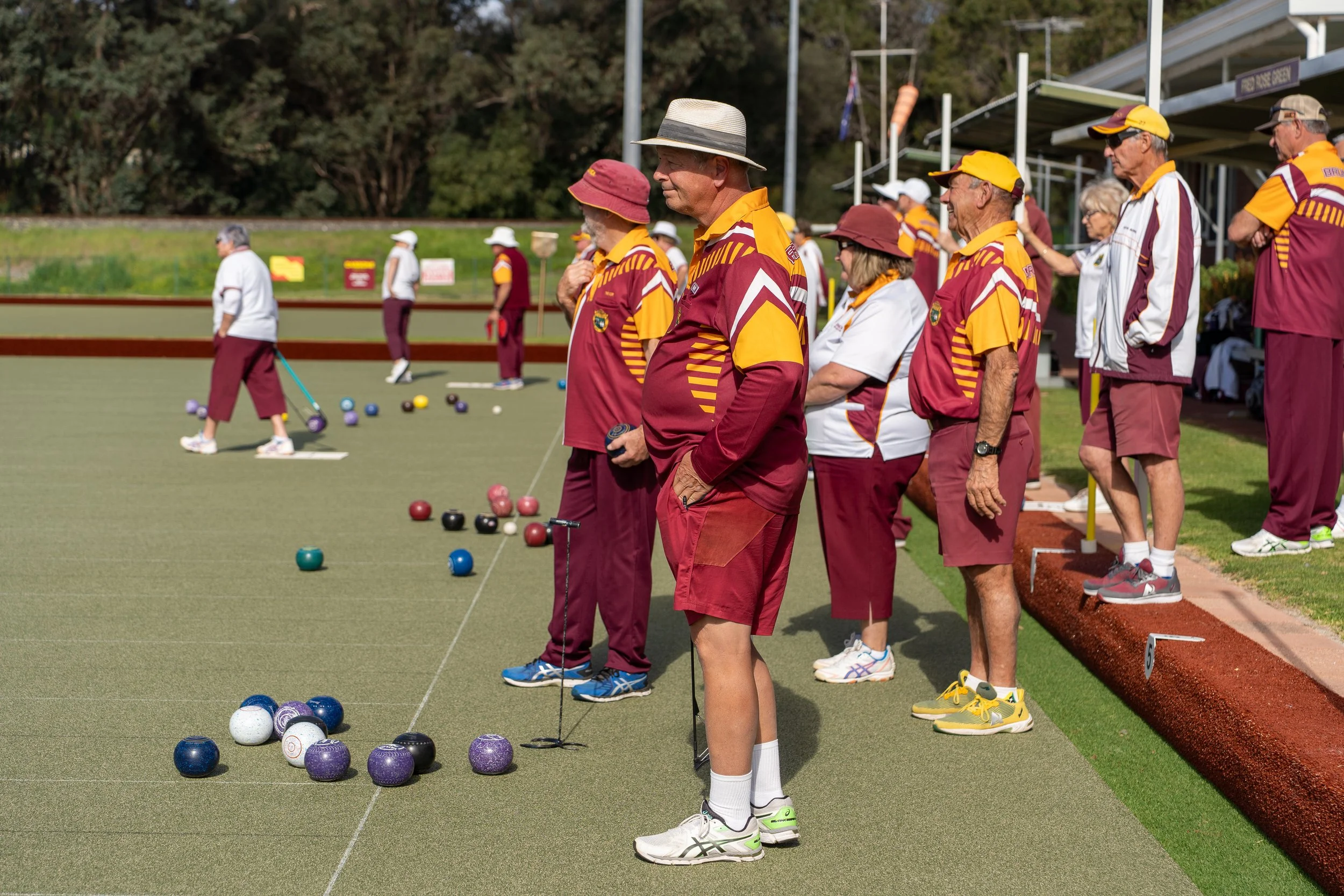 Group of people playing lawn bowls on a green field, wearing sports uniforms, some with hats, surrounded by trees and a stand in the background.