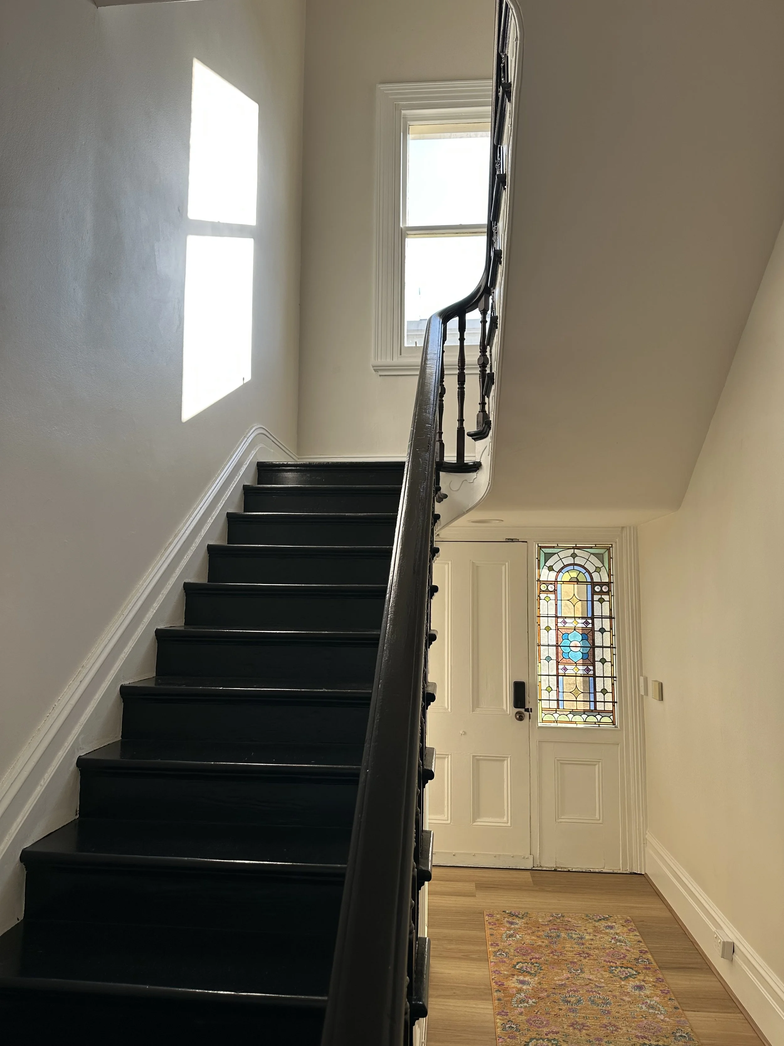 View of a staircase with black steps and a wooden handrail, leading to a door with a stained glass window, with a small colorful rug on a hardwood floor and a window on the wall allowing sunlight to cast a shadow.
